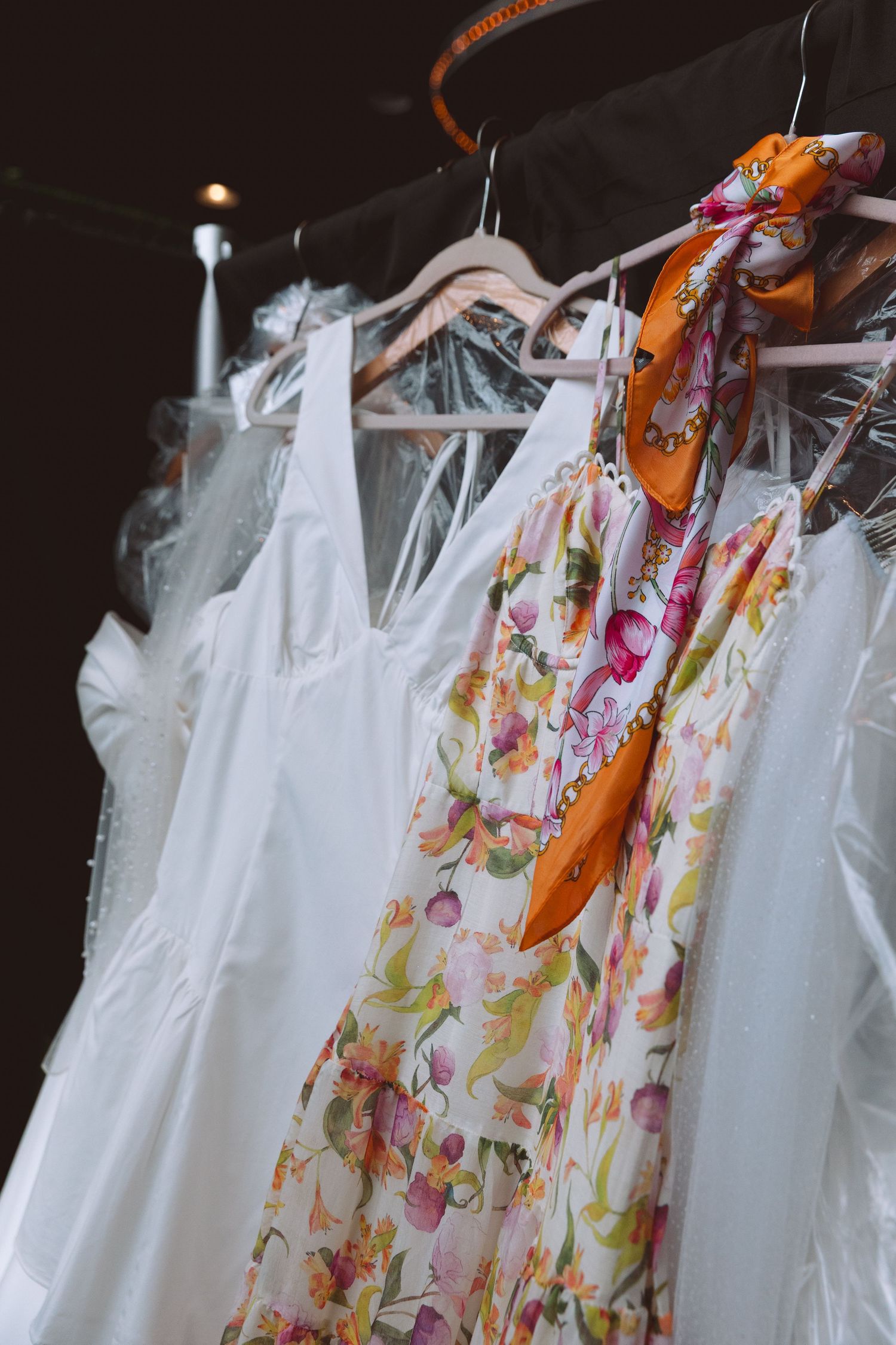 White and floral dresses hanging on a rack with an orange silk scarf draped over a hanger.