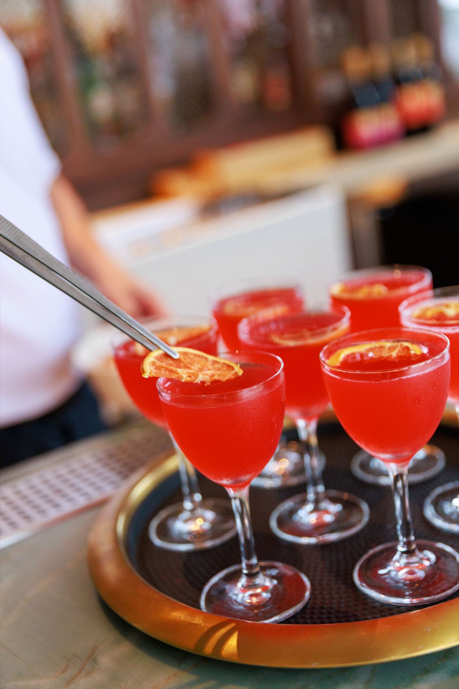 Tray of vibrant red cocktails garnished with dried orange slices, served in elegant stemmed glasses at a bar.