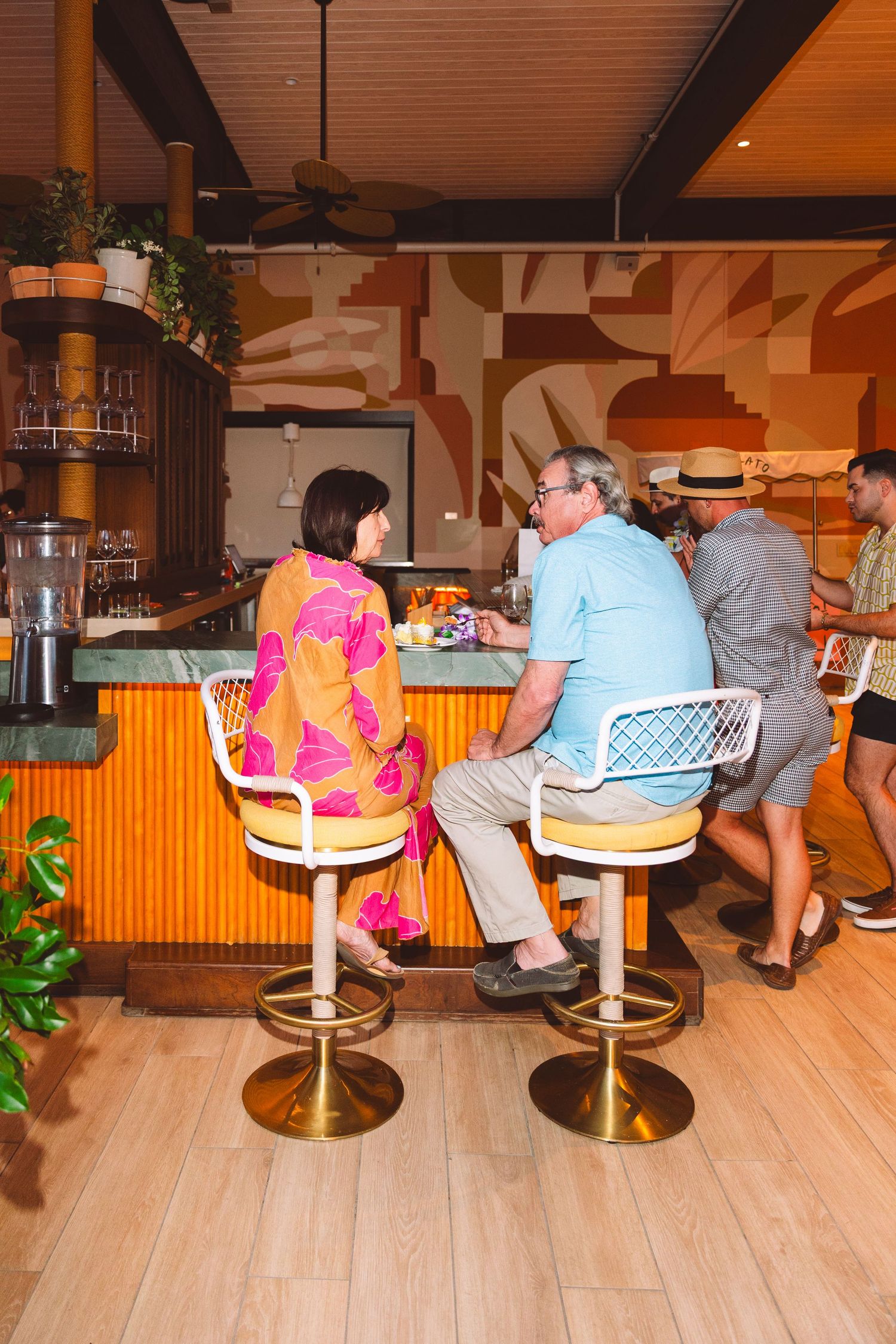 Couple sitting at a colorful retro bar with gold stools, tropical decor, and a vibrant geometric mural backdrop.