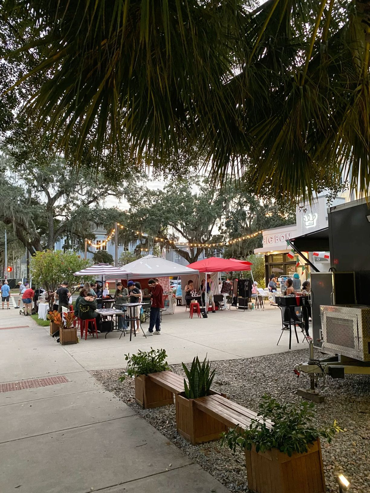 Outdoor food market with colorful vendor tents, string lights, and palm trees in a vibrant urban plaza.