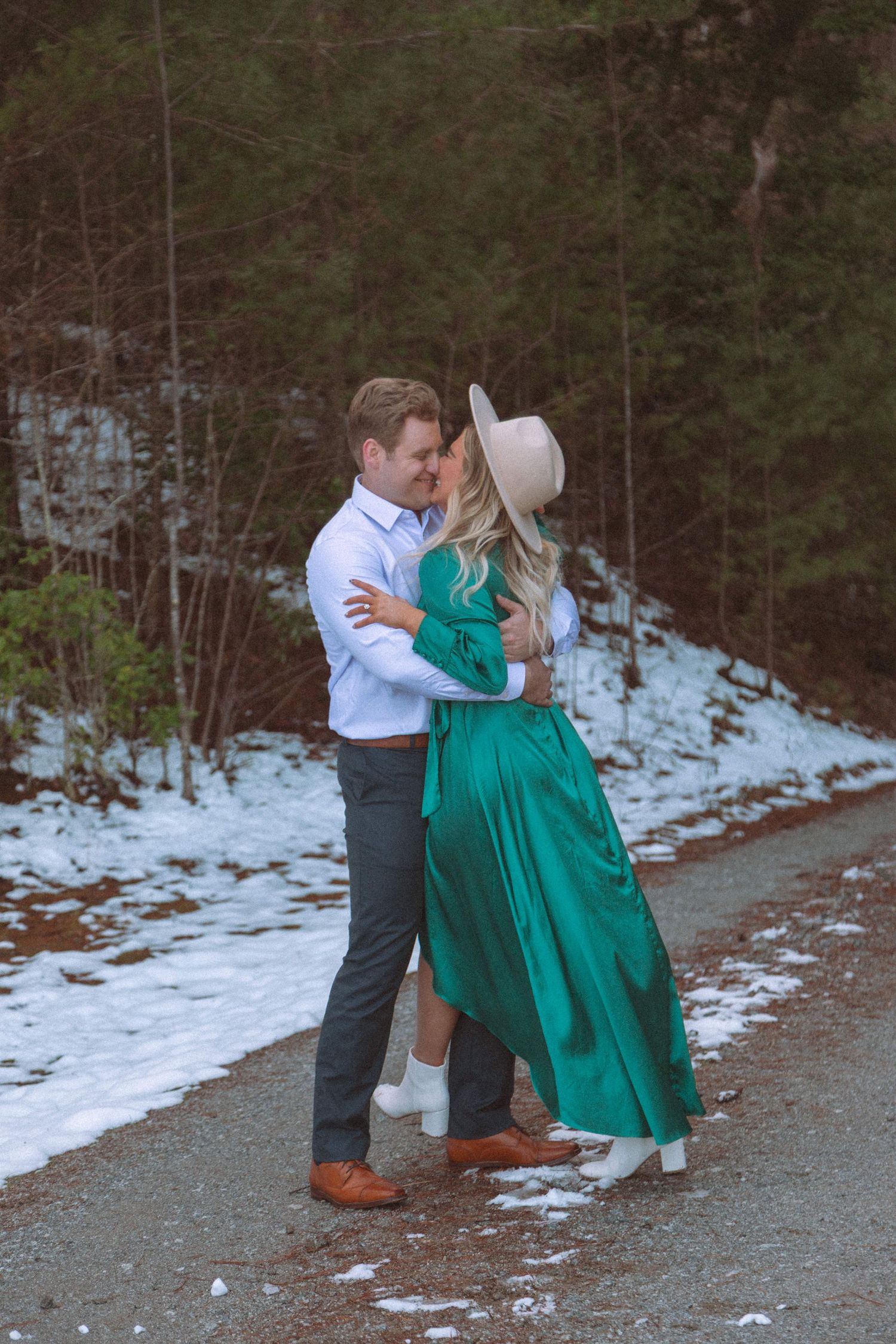 Couple embracing on a snowy road surrounded by winter forest, woman wearing a green dress and hat.