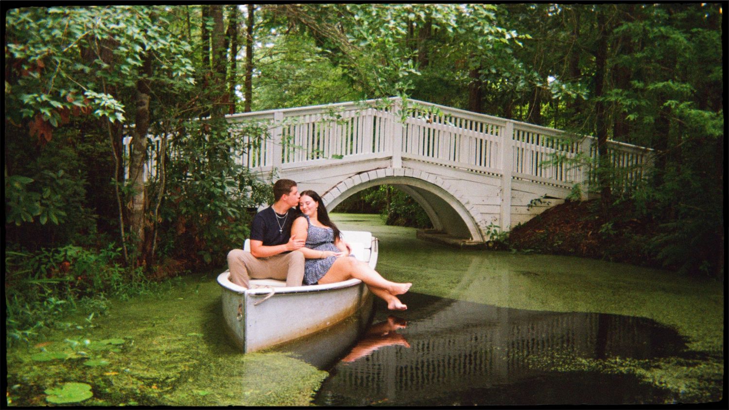 Couple kissing in a white rowboat on a serene green pond beneath a charming white arched bridge.