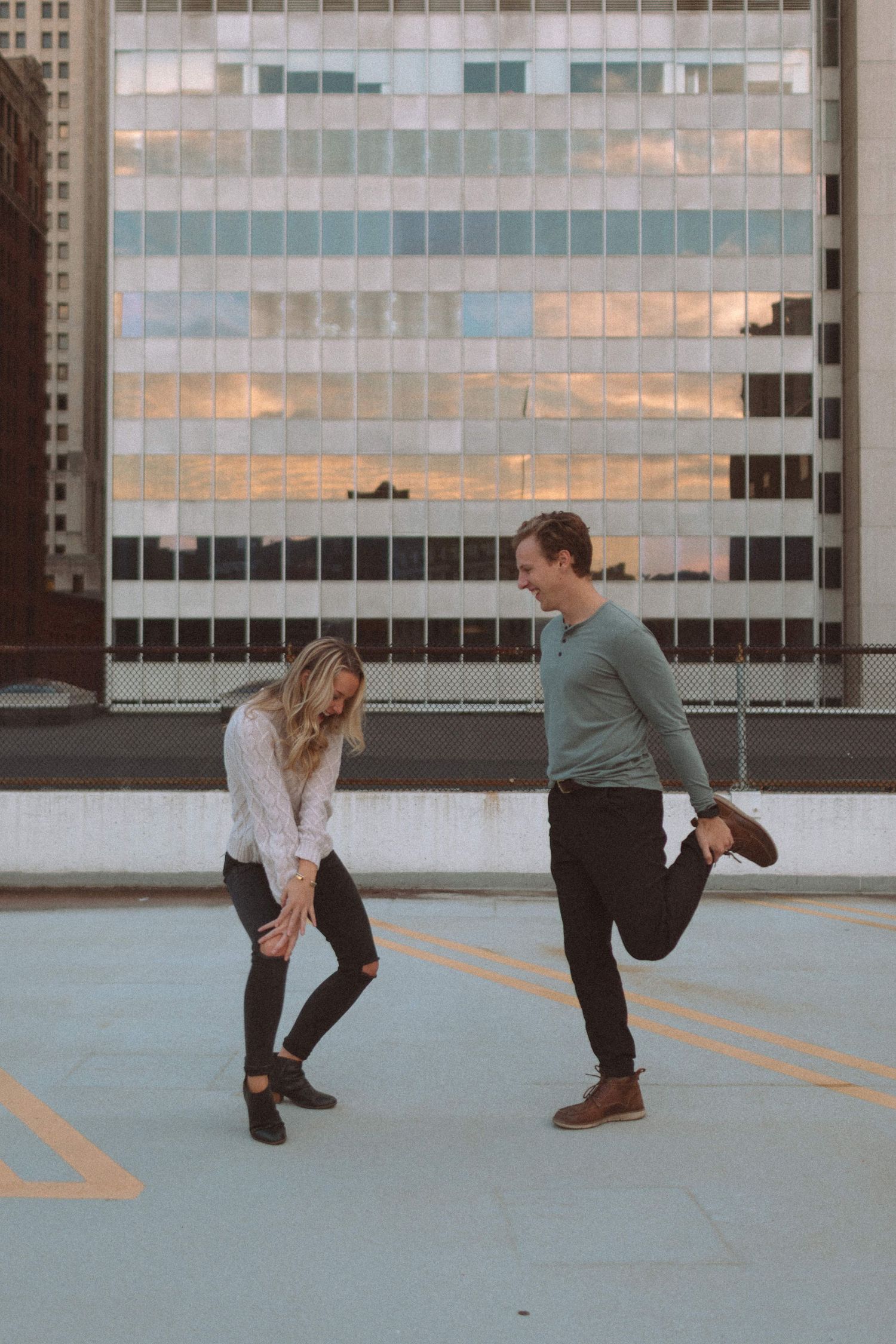 Laughing couple playfully kicking legs on rooftop parking with city building reflected at sunset.