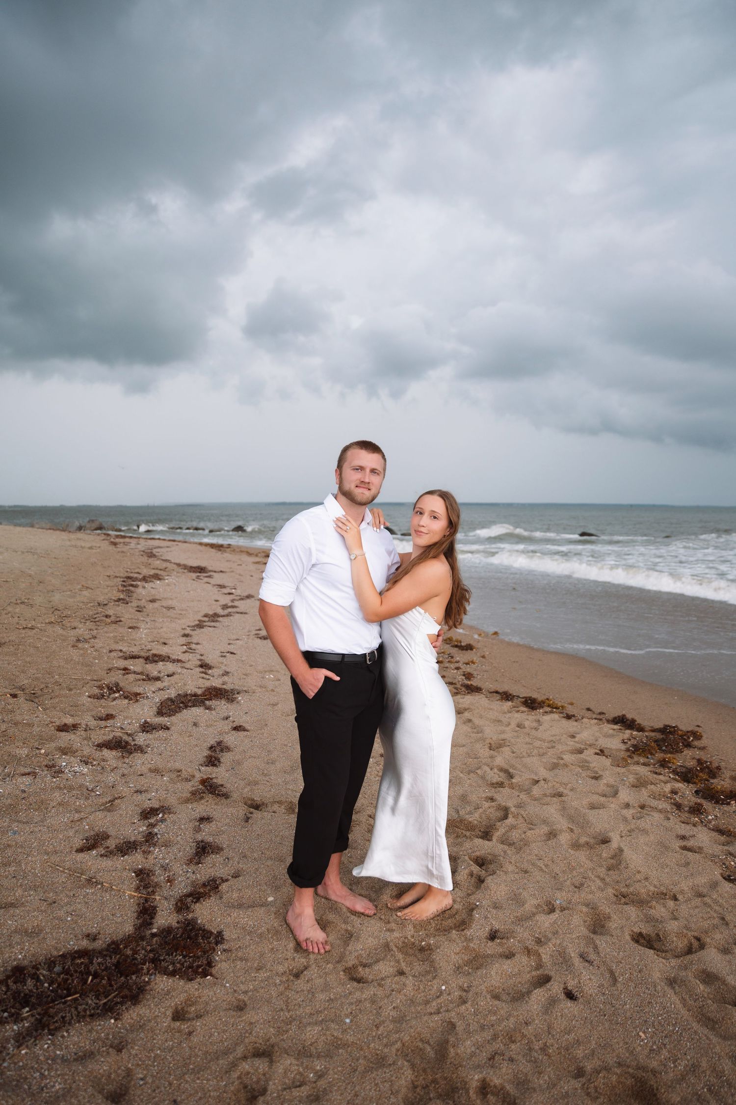 Couple embracing on a sandy beach with stormy ocean waves and dramatic cloudy sky behind them.