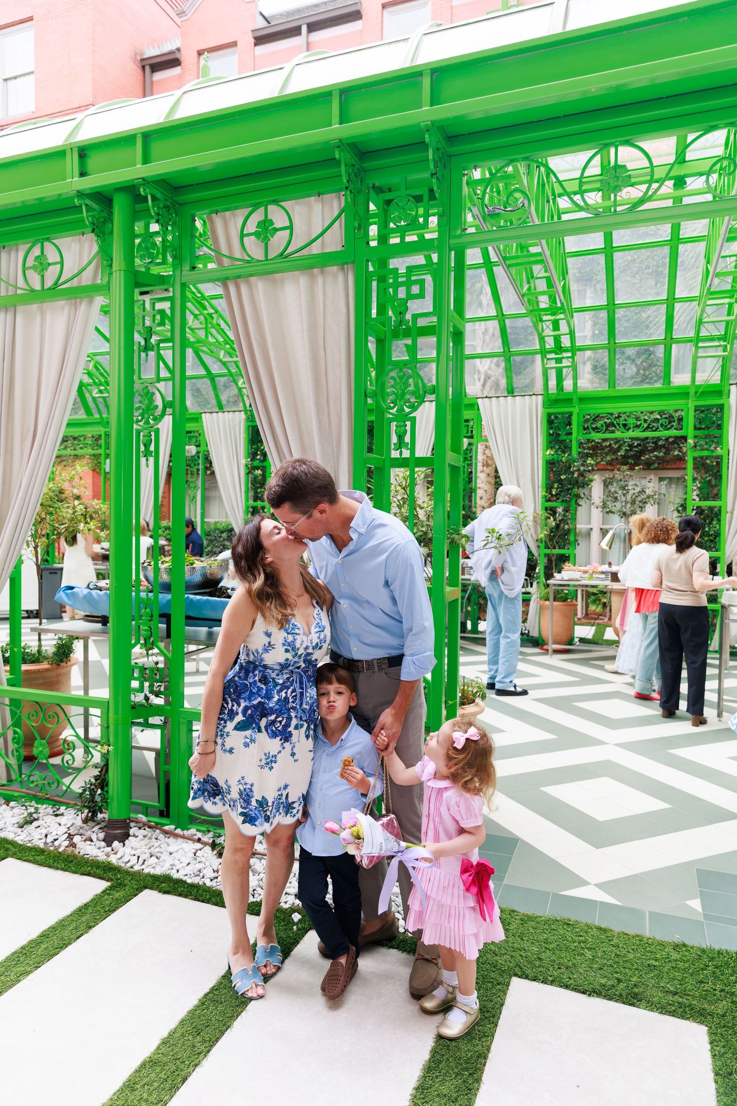 A couple kisses while their two young children stand beside them in a vibrant green outdoor pavilion.