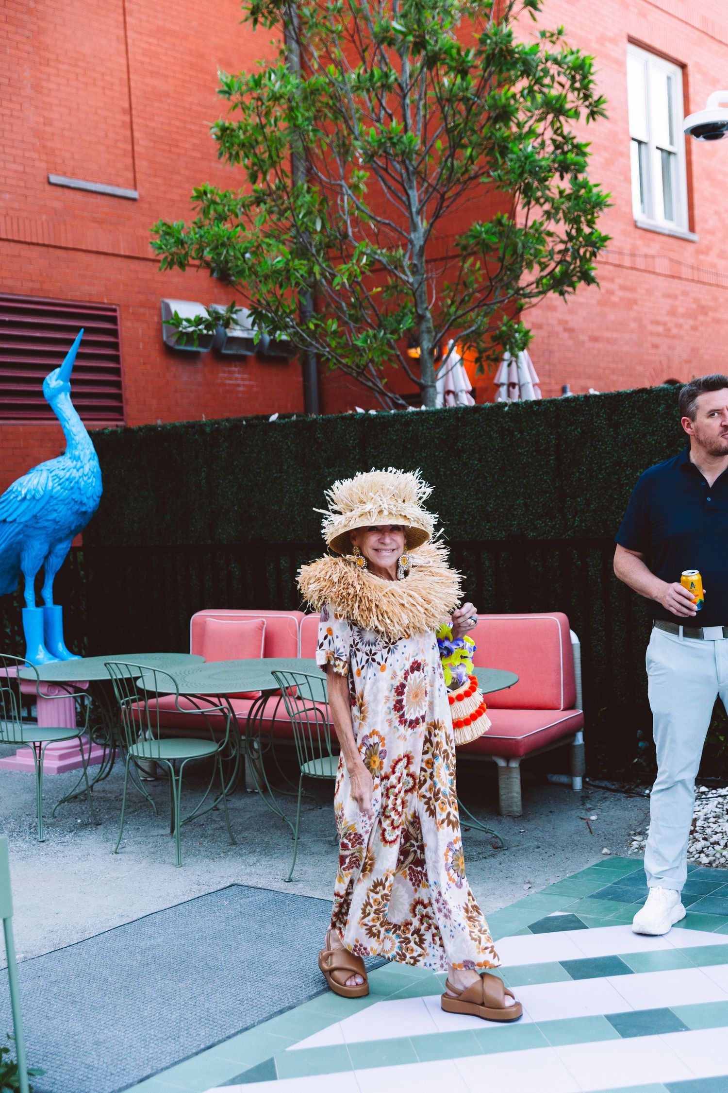 Stylish woman in floral wide-leg pants and straw hat poses in colorful outdoor patio with blue heron sculpture.