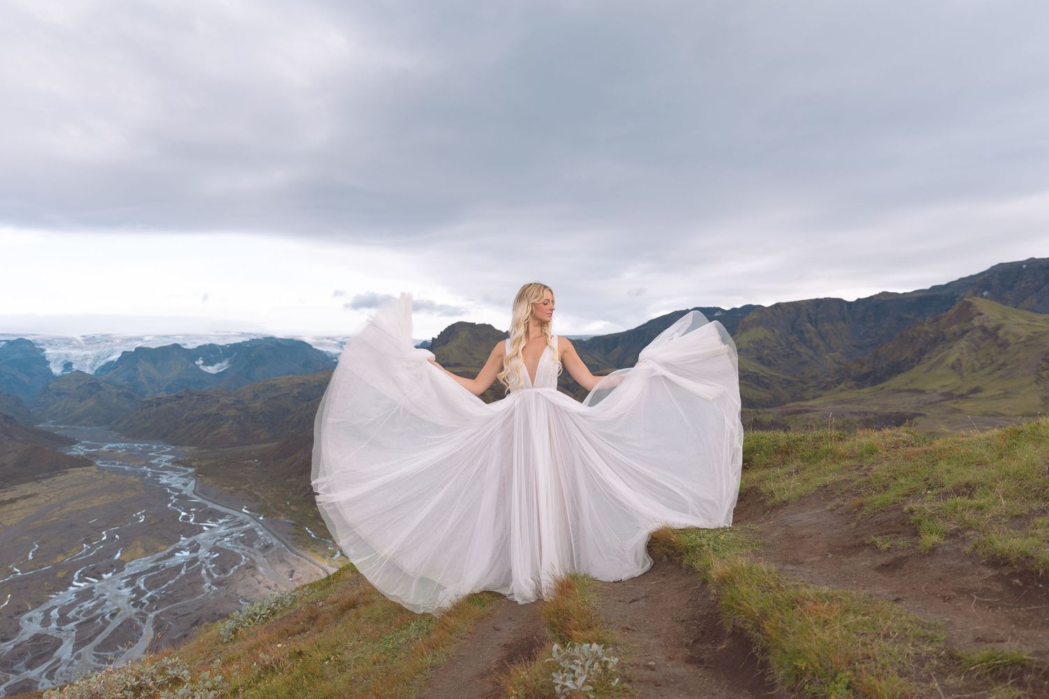 Bride in flowing white gown spreading her dress on a scenic mountain overlook with dramatic cloudy skies in Iceland.