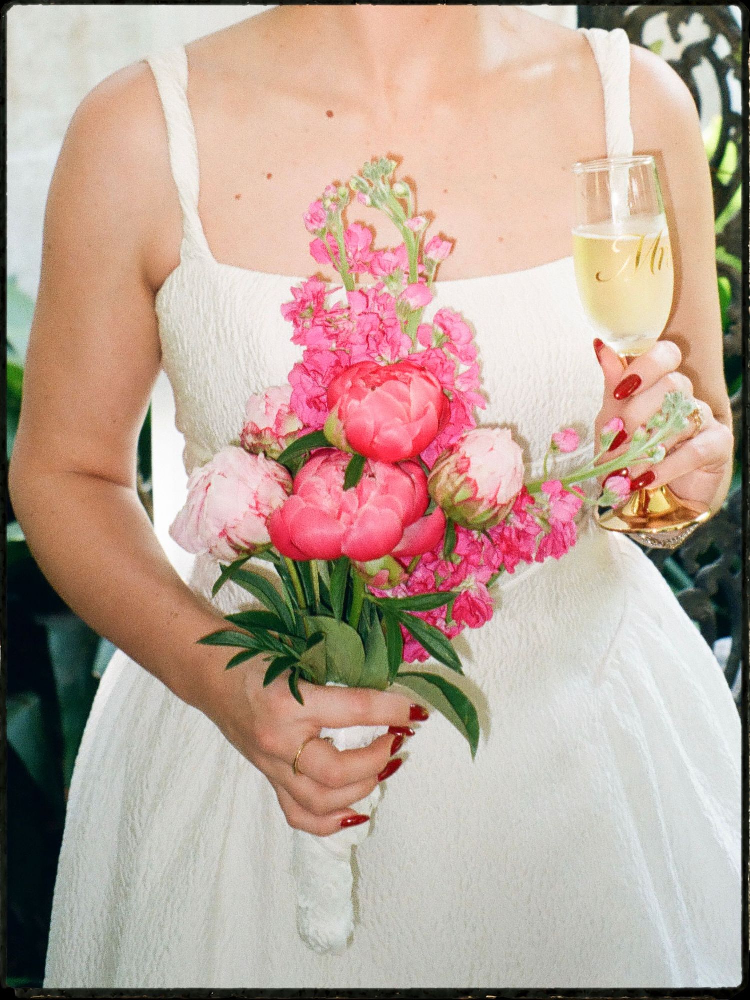 Bride holding a vibrant pink peony and snapdragon bouquet with a glass of champagne in a white dress.
