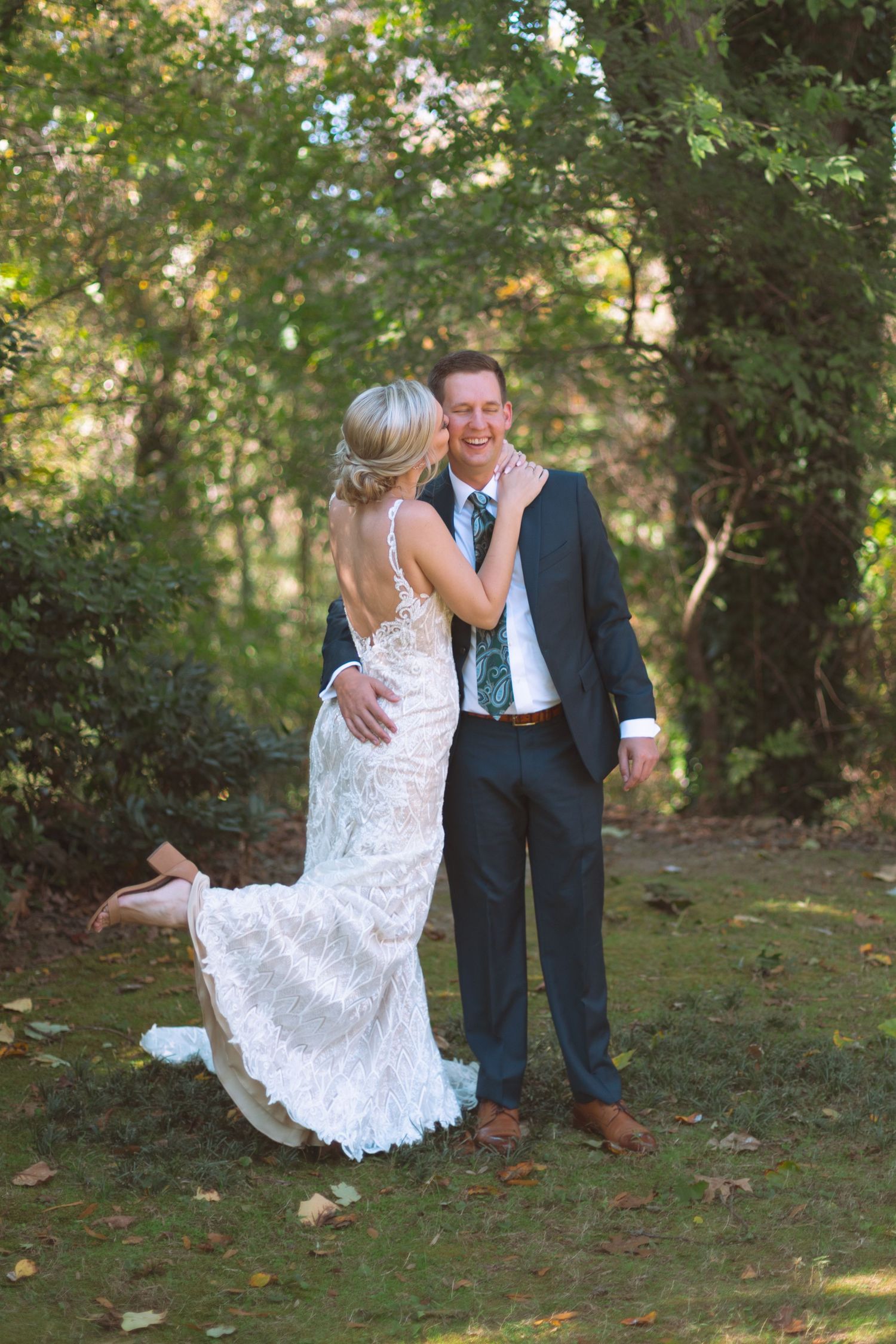 Joyful bride in lace gown kisses smiling groom during outdoor first look moment among green trees.