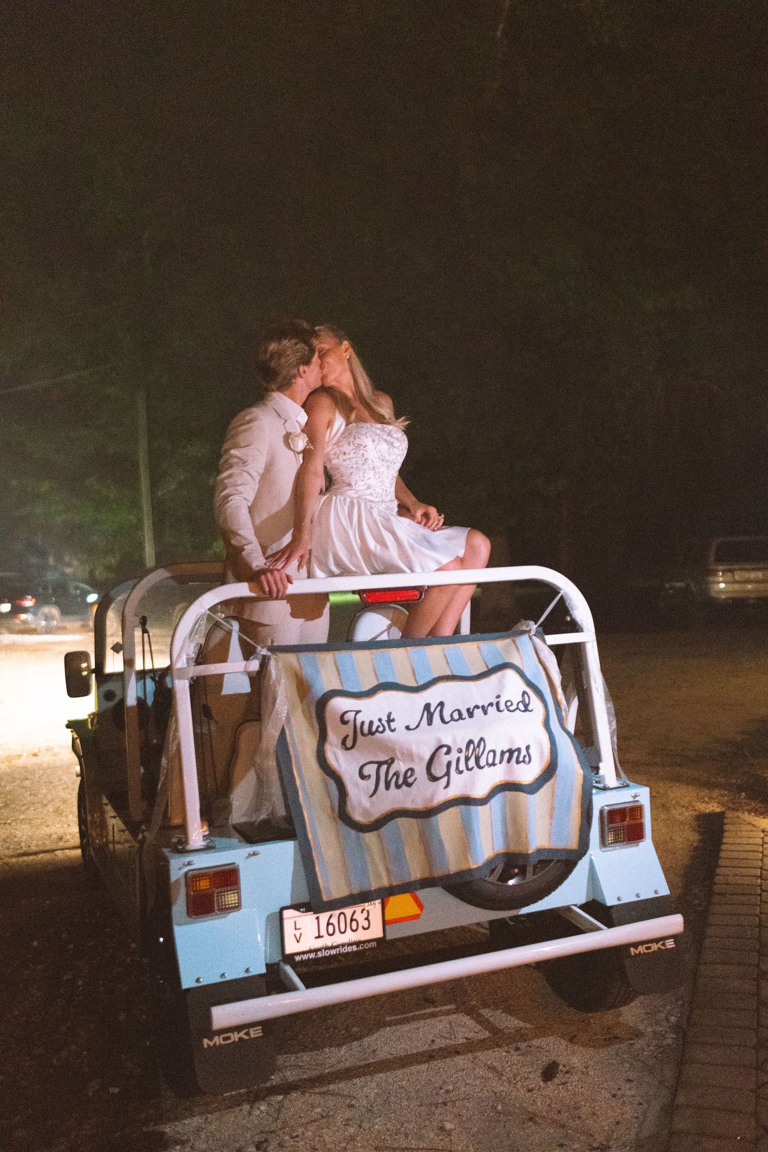 Just married couple kissing on back of vintage blue golf cart with Just Married The Gillams sign at night.