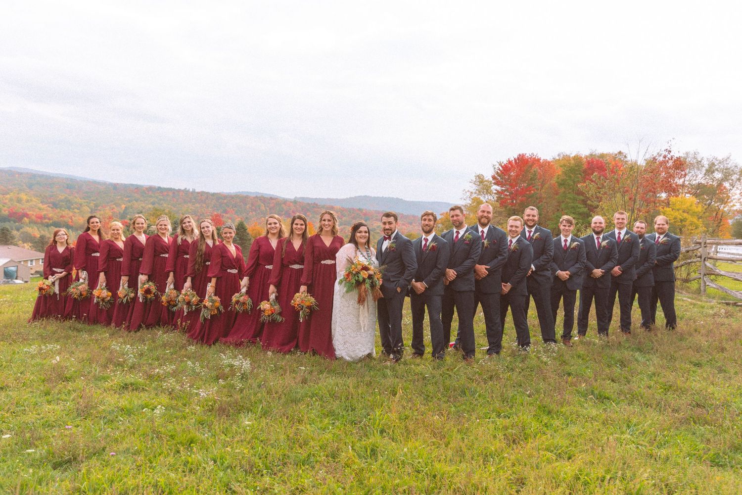 Large wedding party poses outdoors with bridesmaids in burgundy gowns and groomsmen in gray suits against colorful fall foliage.