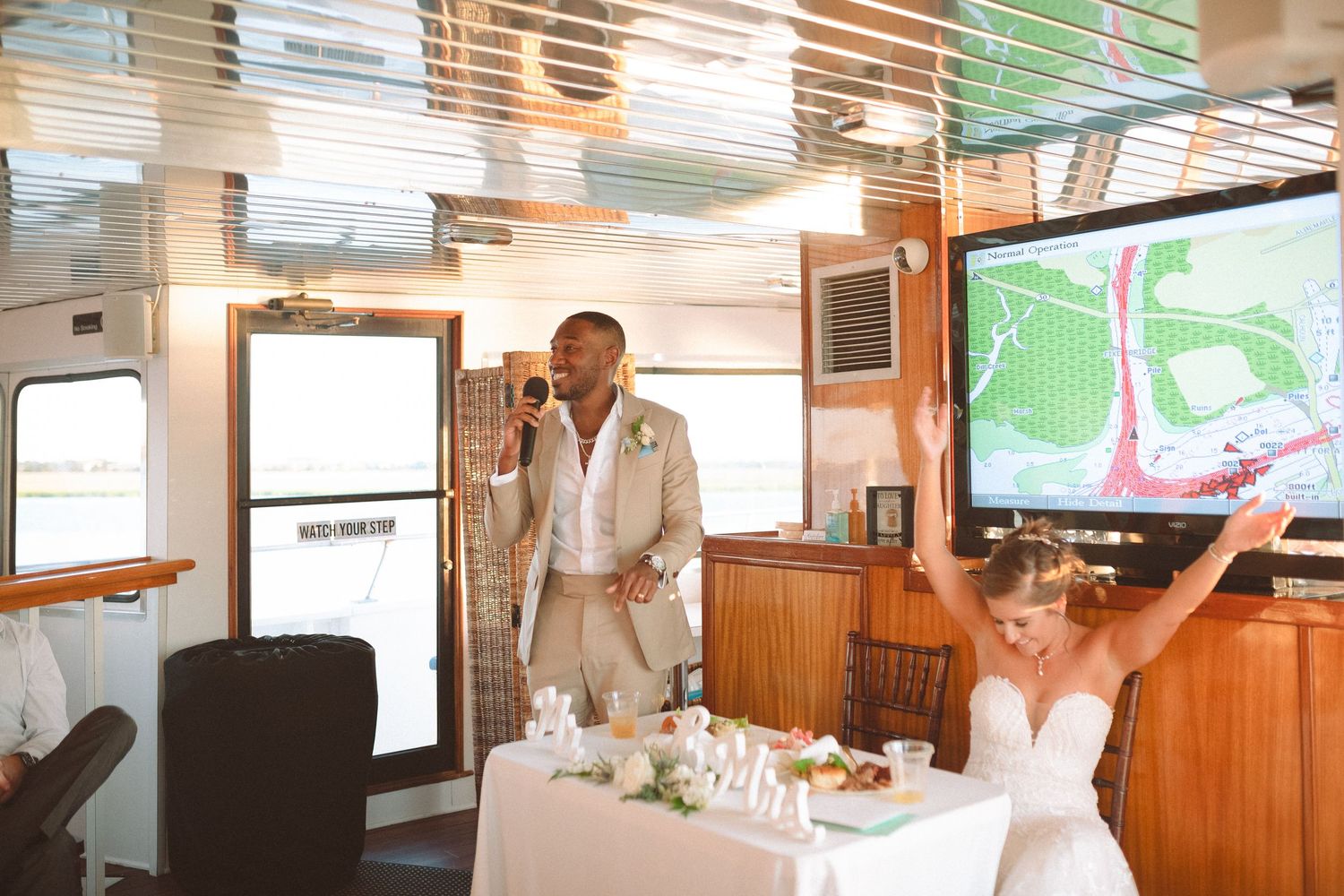 Groom delivers speech as bride raises arms joyfully at decorated sweetheart table on yacht.