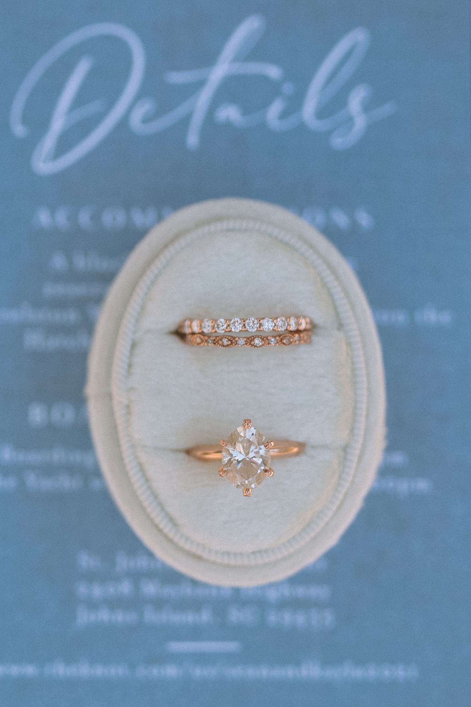 Two rose gold diamond wedding rings displayed in oval velvet ring box on blue wedding stationery.
