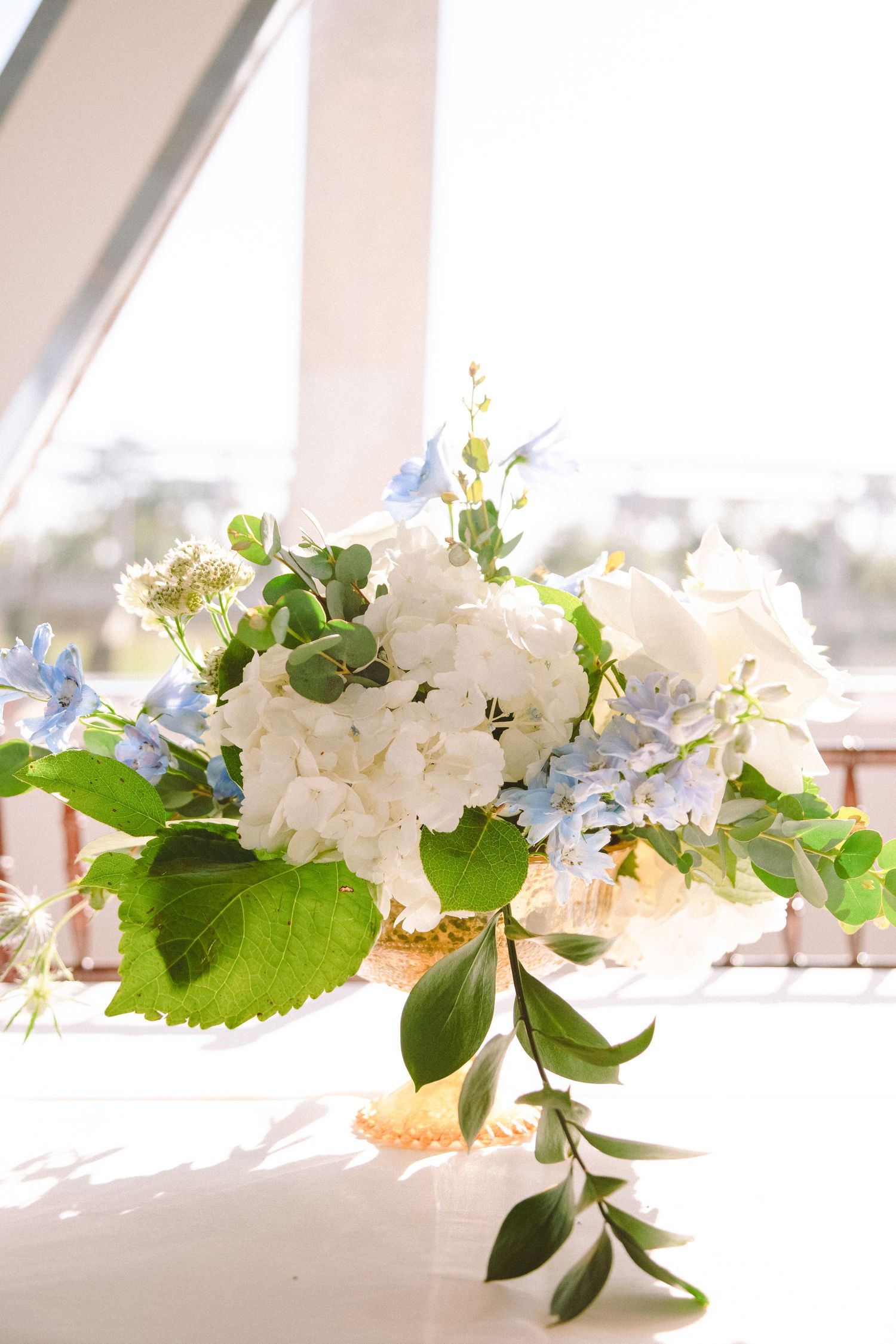 A lush bridal centerpiece of white hydrangeas, blue flowers, and greenery in a gold vase by a window.