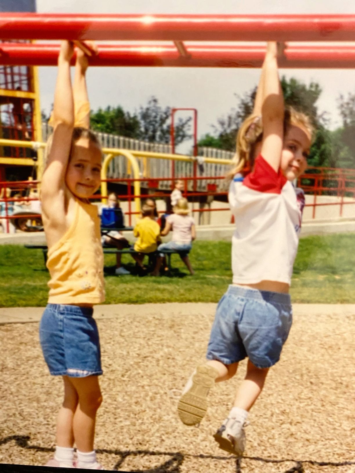 Two children hang from monkey bars at a sunny outdoor playground.