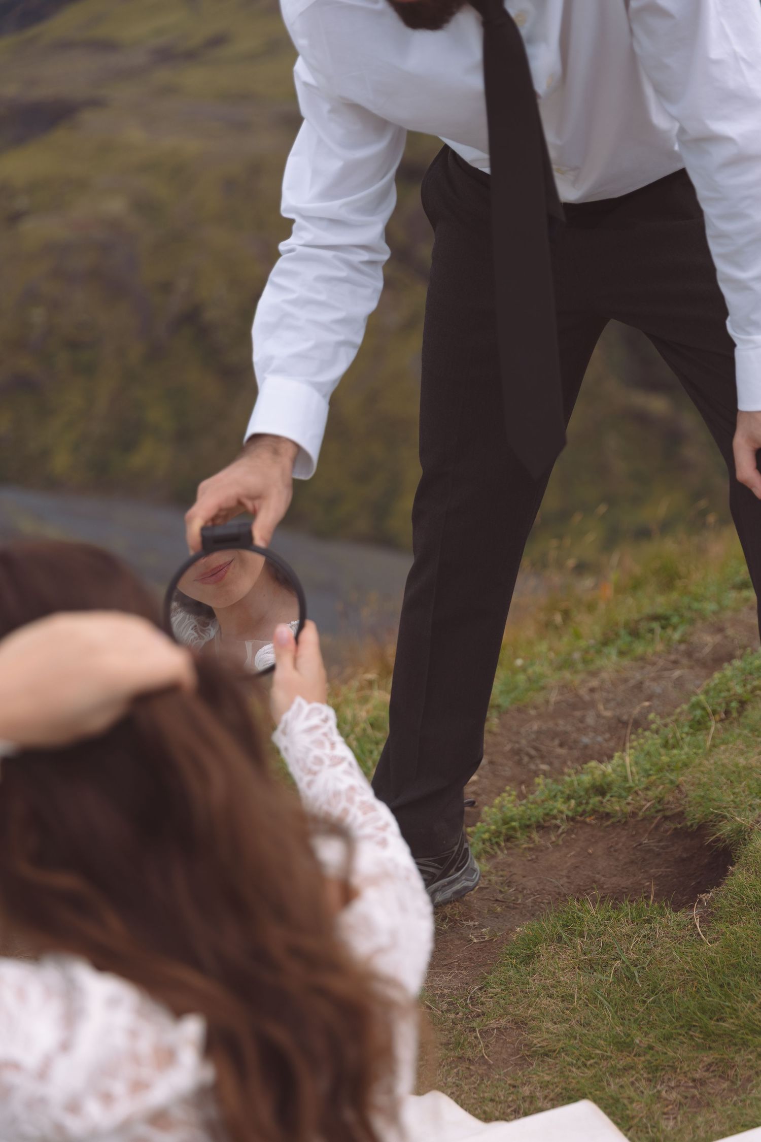 Wedding couple examining something with magnifying glass on grassy hillside in formal attire.