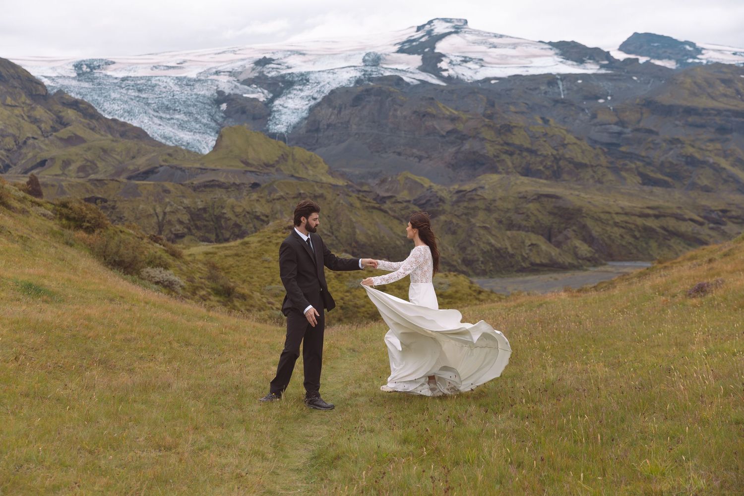 Bride in flowing white dress and groom dancing on green meadow with glacier backdrop.