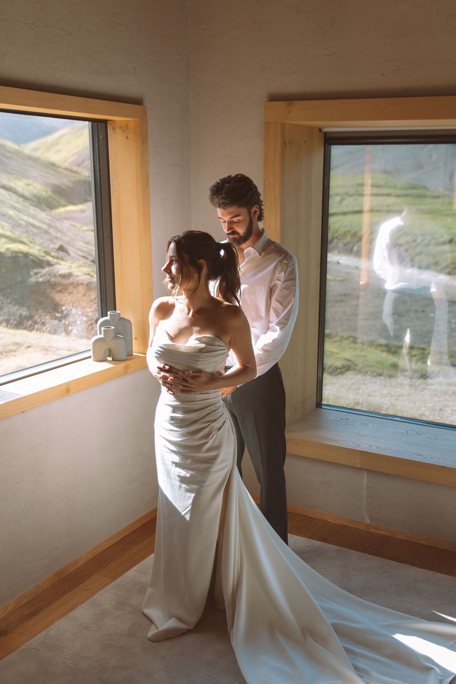 Groom tenderly embraces bride in white satin gown by sunlit windows with mountain views.