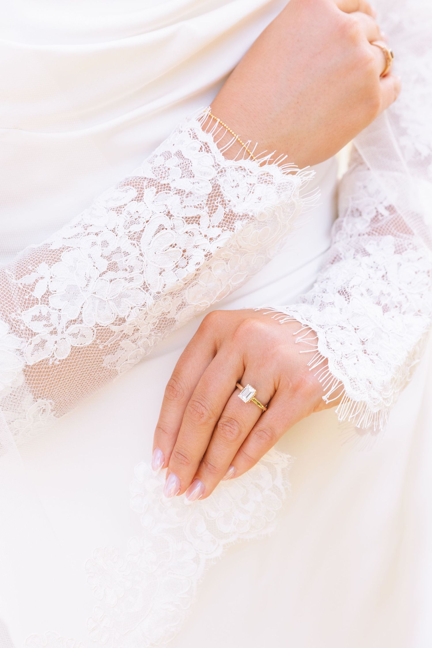 Close-up of bride's hand with emerald cut diamond engagement ring on lace wedding dress sleeve.