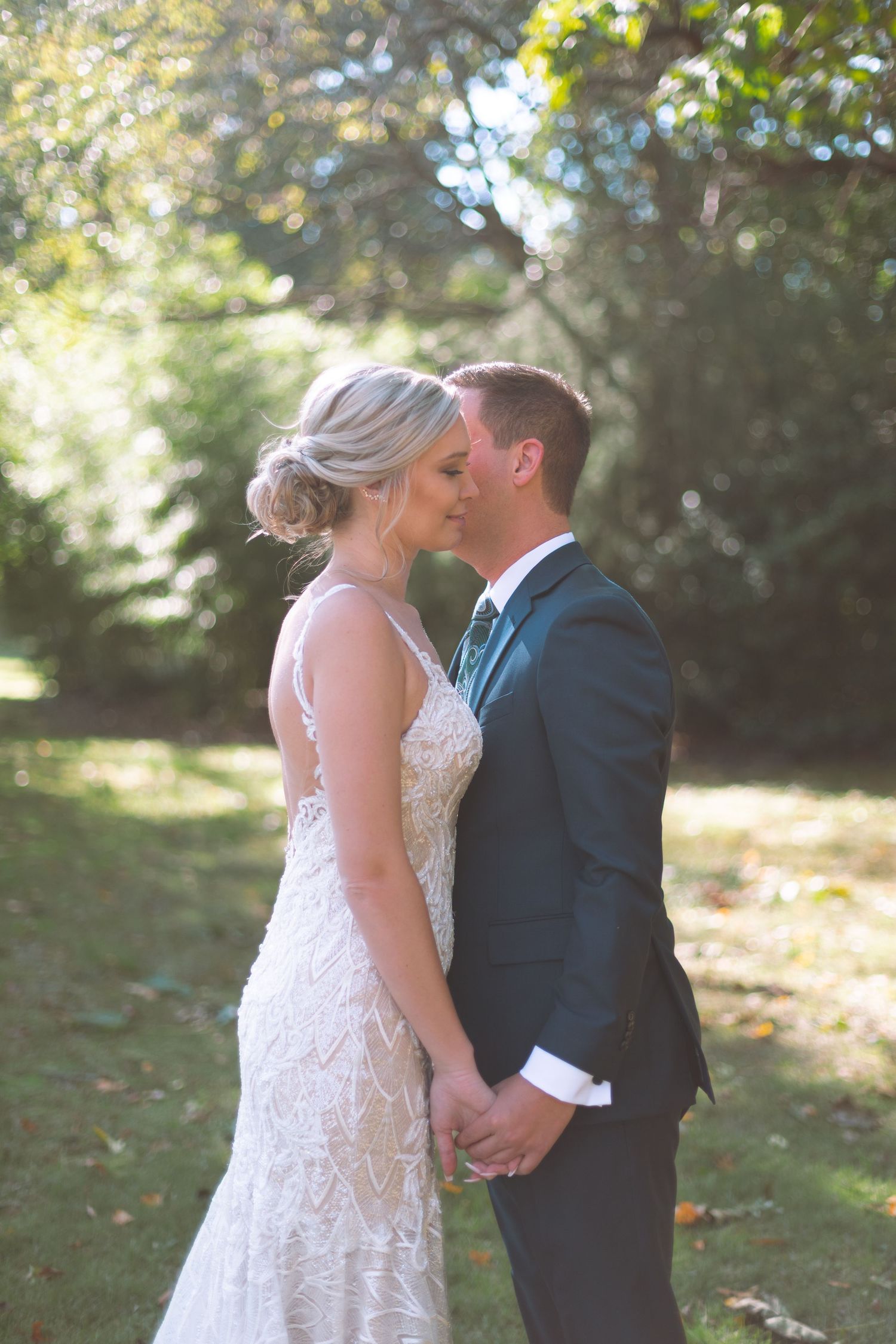 Groom kissing bride on forehead while holding hands in a sunlit garden setting.