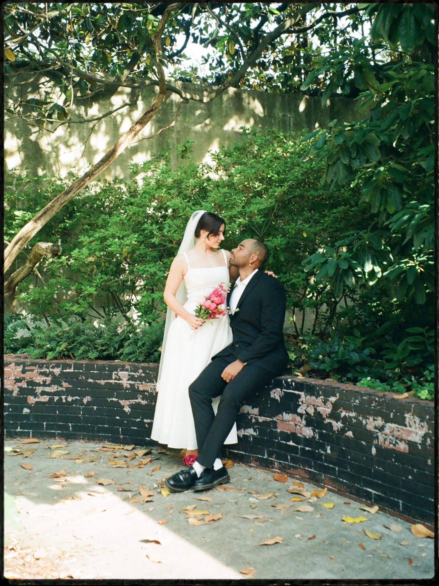 Interracial couple shares tender kiss near brick wall surrounded by lush green garden foliage.