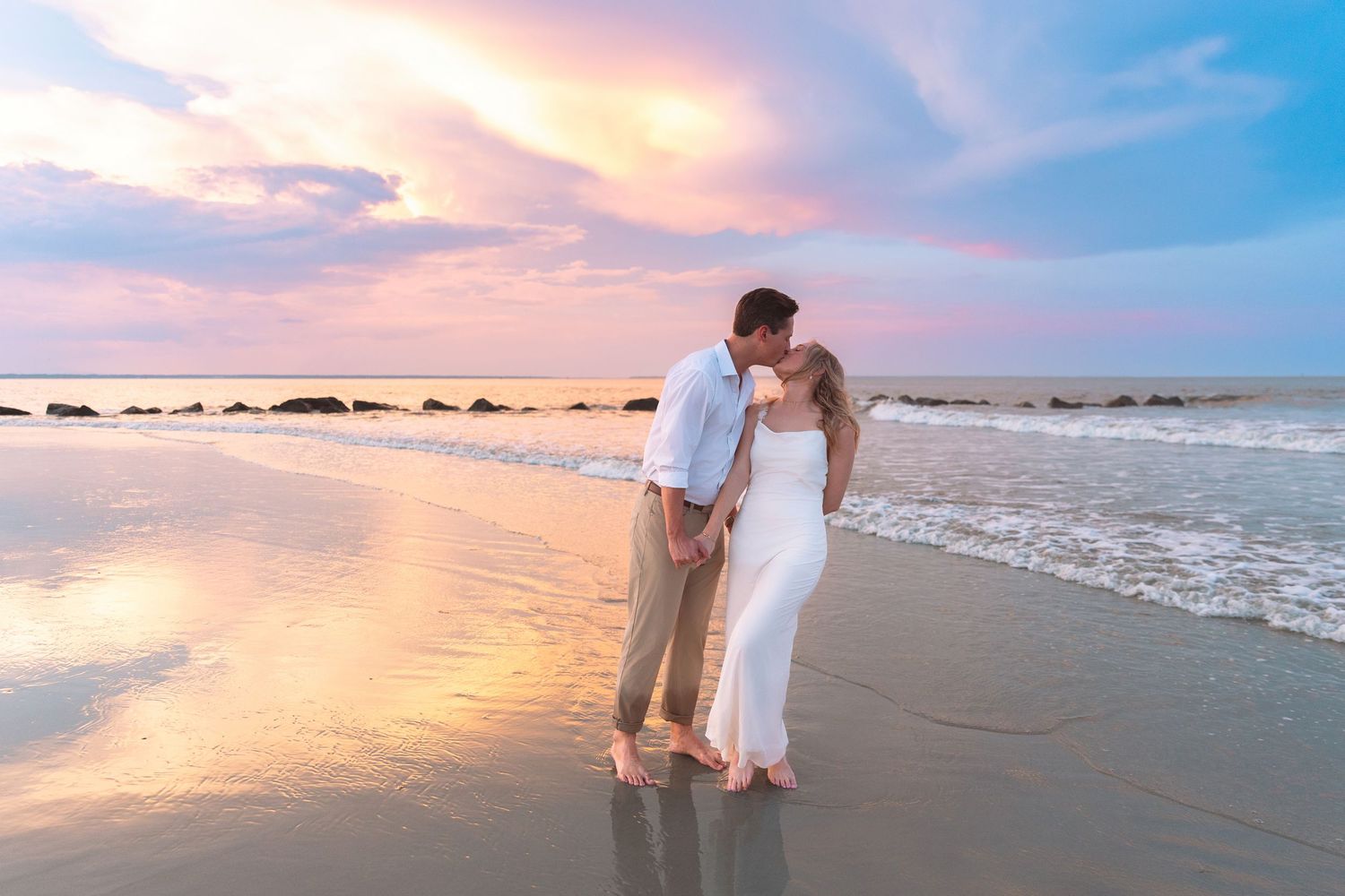 Couple kissing on a beach at sunset with colorful pink and purple sky reflection.