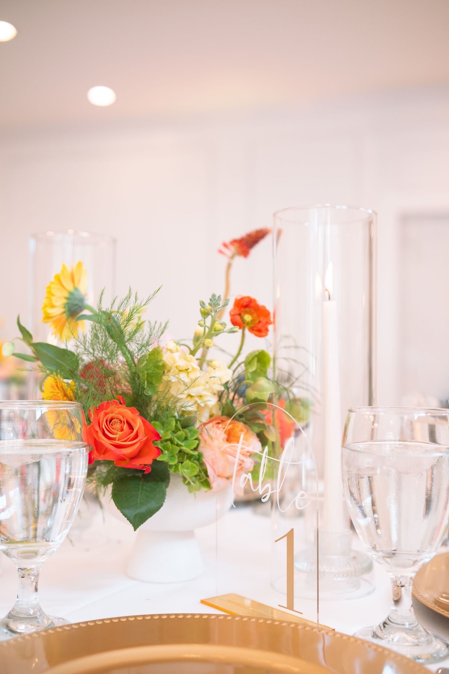 Wedding reception table setting with colorful floral centerpiece, crystal glasses, and gold charger plates.