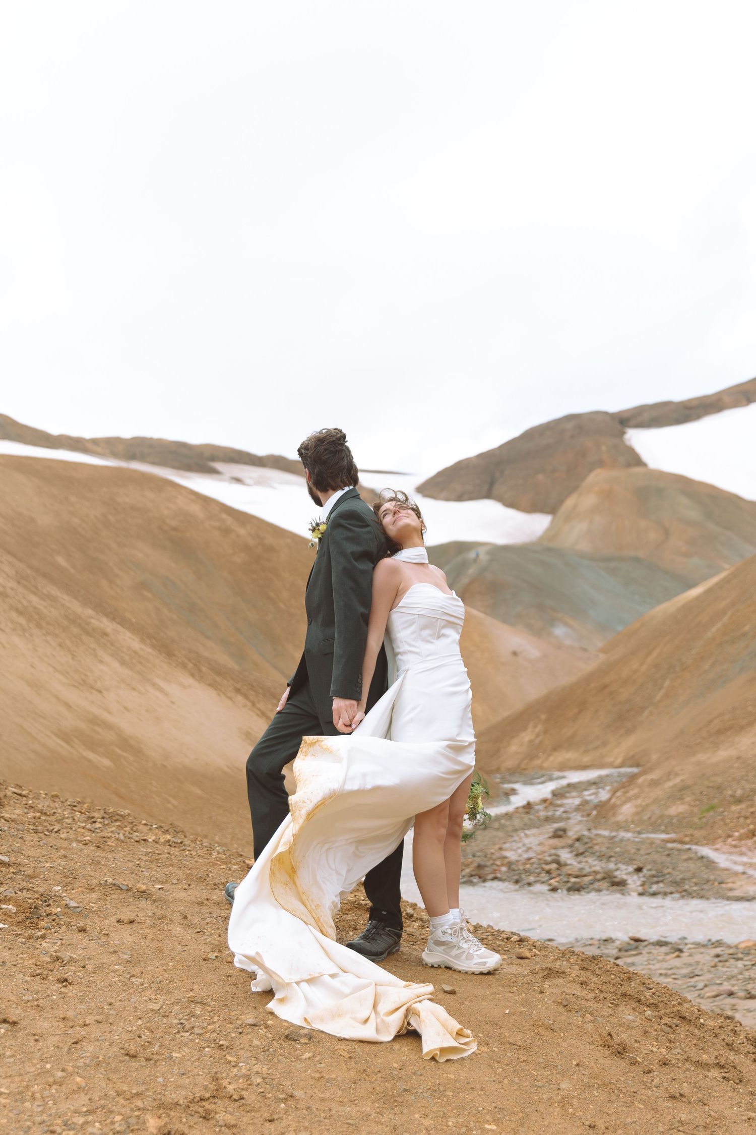 Bride and groom embrace on rocky mountain terrain, her white gown flowing in the wind during an outdoor elopement.