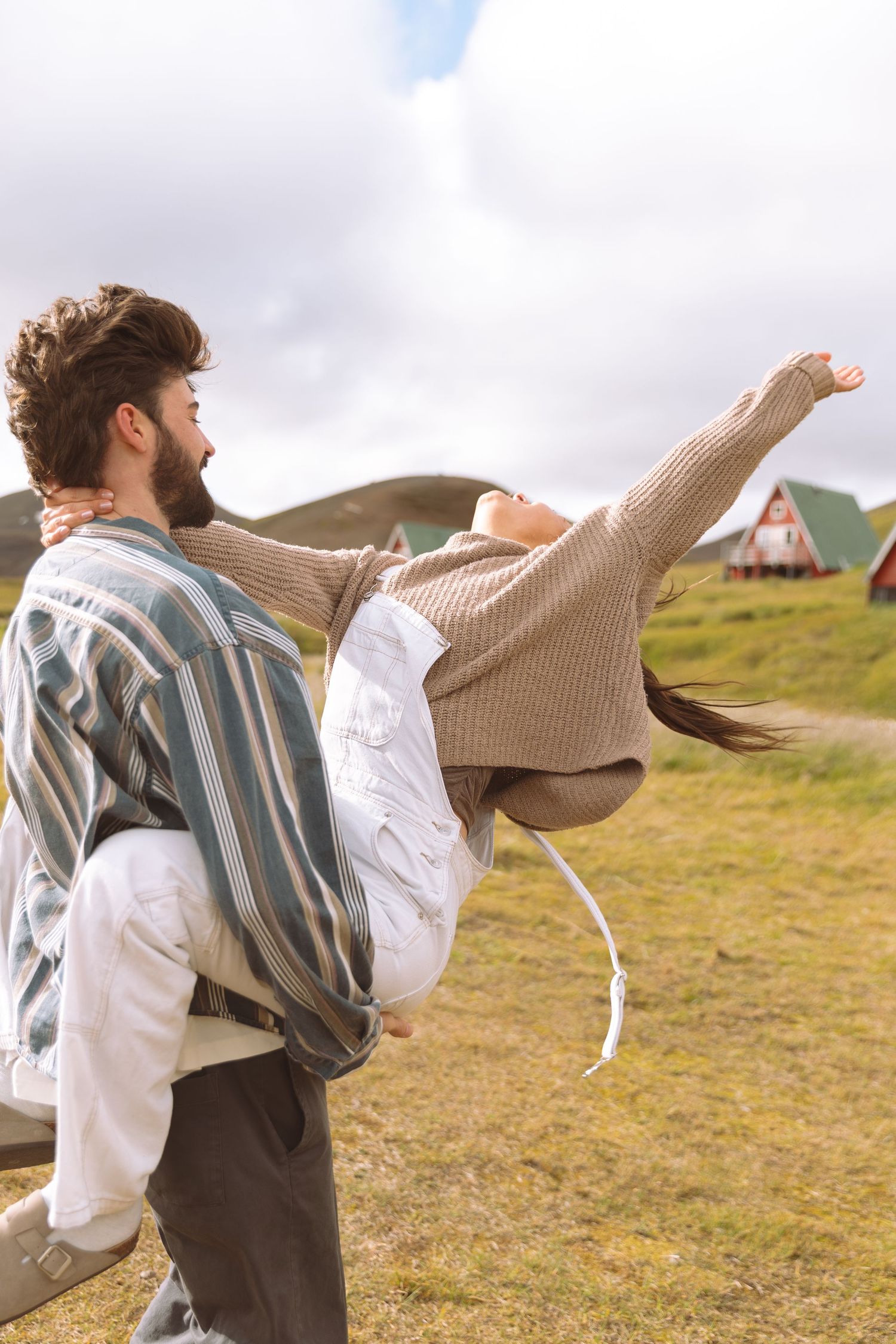 Man carrying woman on his back in a green valley with colorful cabins visible in the distance.