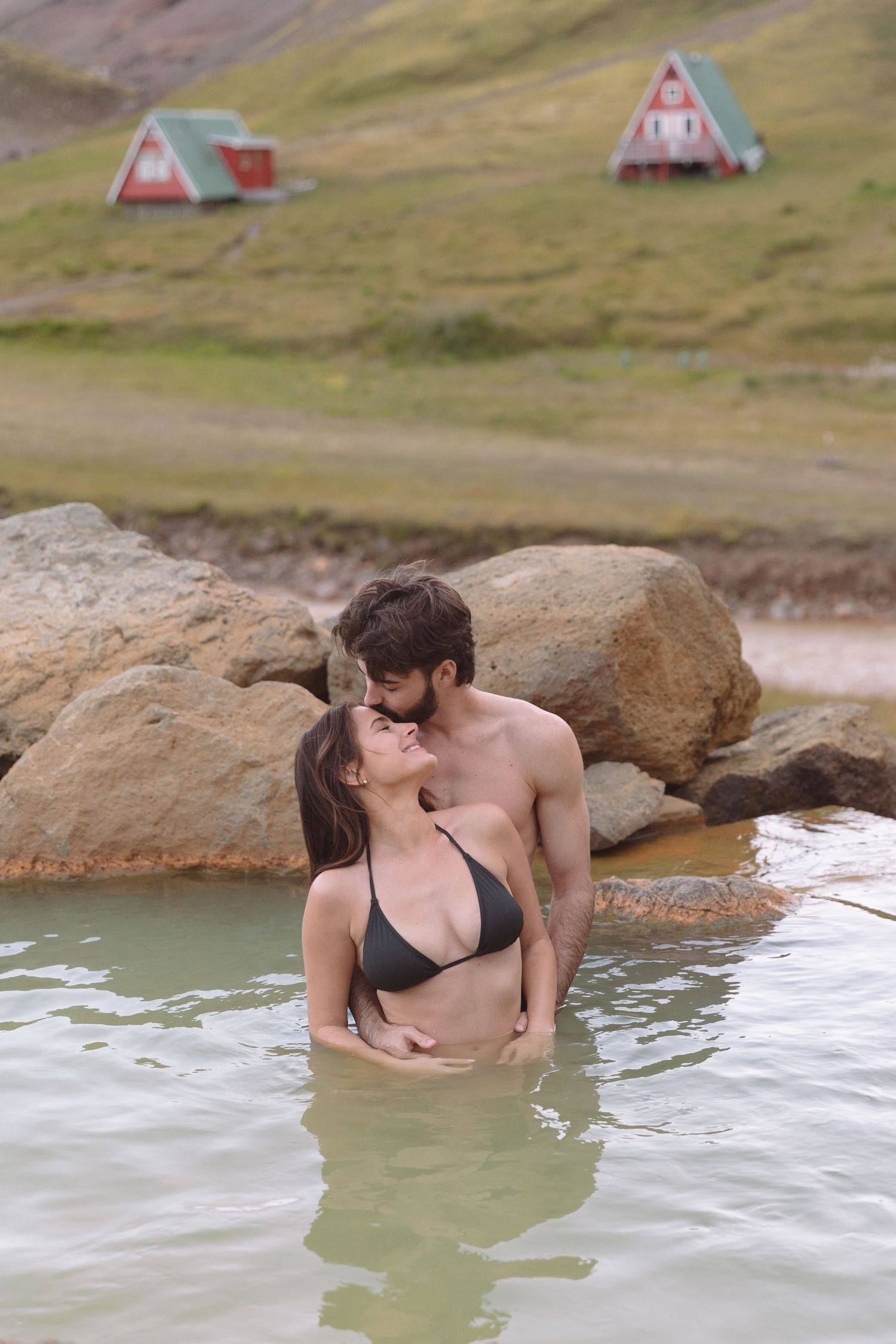 Couple sharing a kiss in a natural hot spring pool with scenic Icelandic hillside cabins behind them.