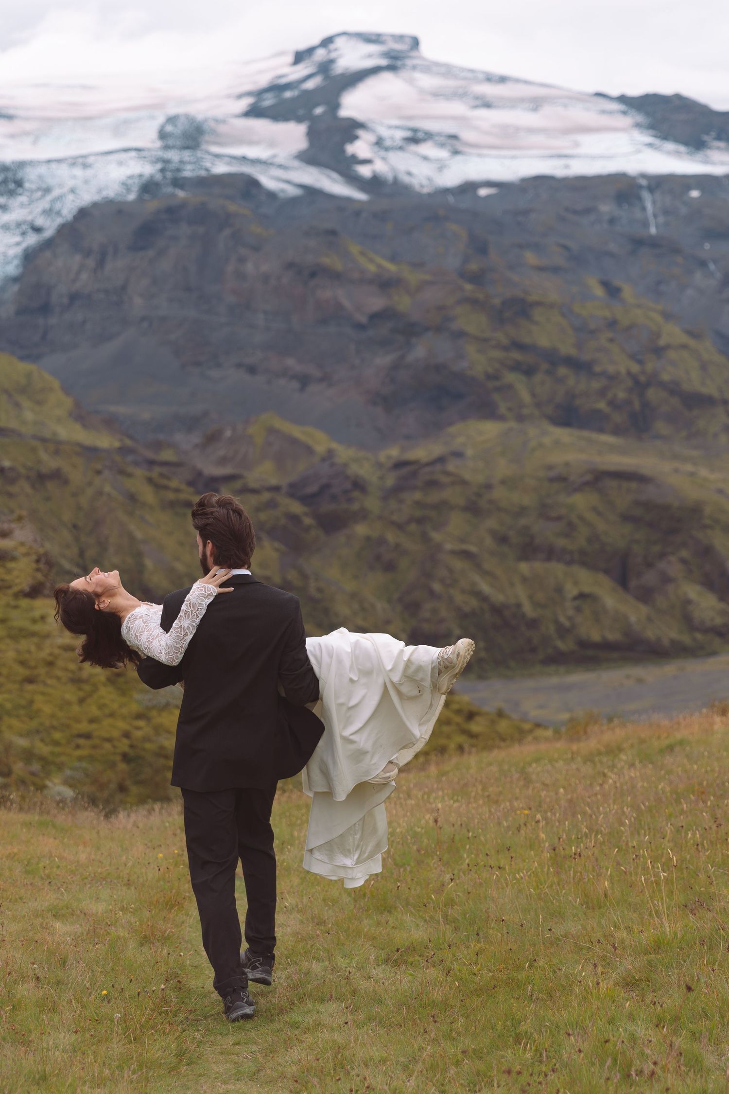 Groom carries bride through an open meadow as she leans back with arms outstretched near a glacier.