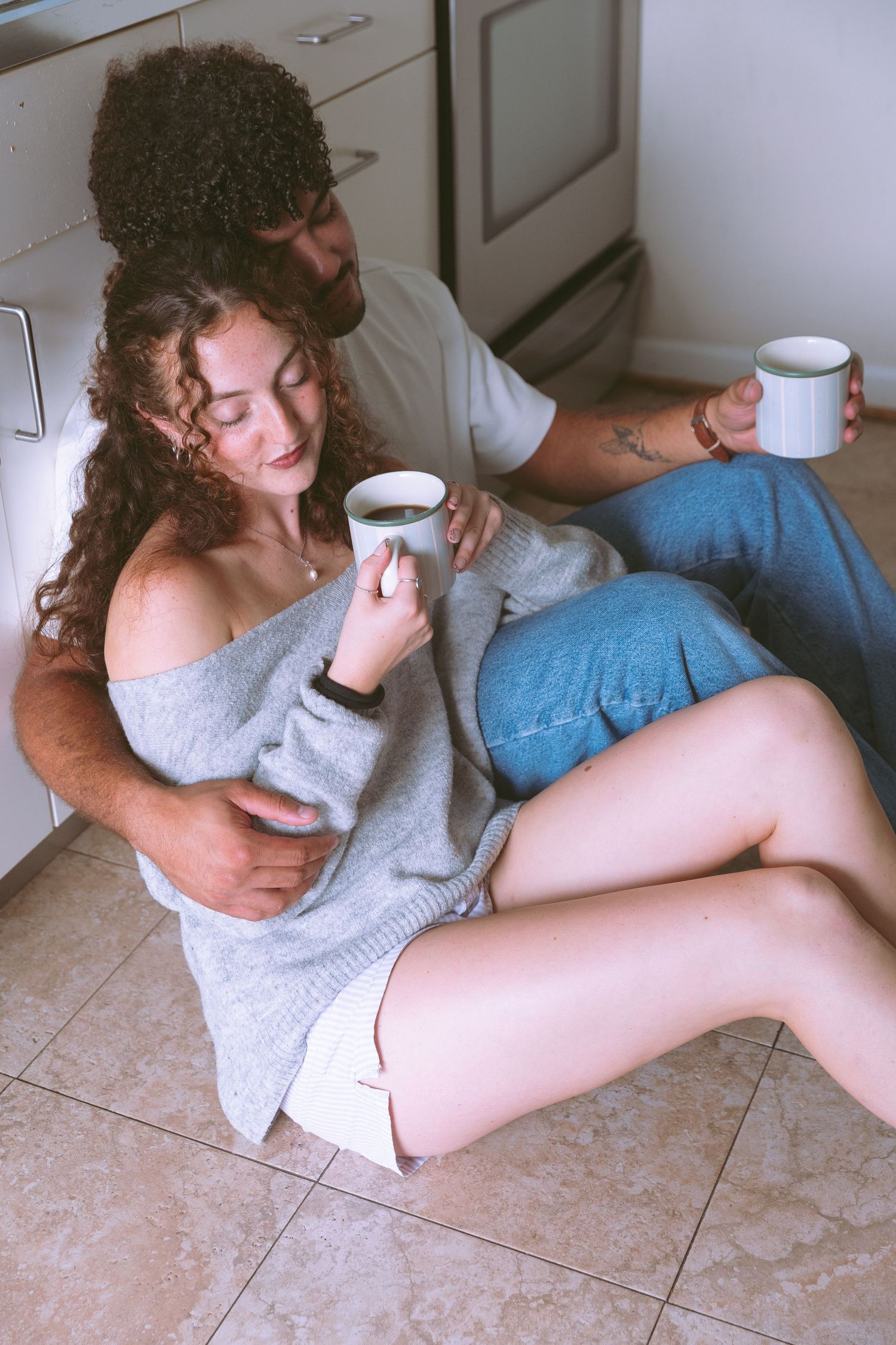 Young couple sitting on kitchen floor together sharing mugs of coffee.