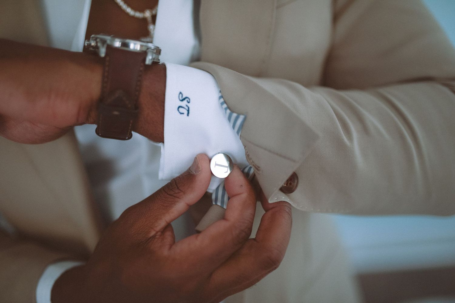 Close-up of groom's monogrammed cufflinks being fastened on white dress shirt with beige wedding suit.
