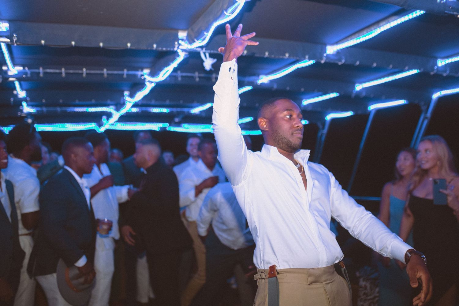 Groom raising arm on crowded wedding reception dance floor under blue neon lights.