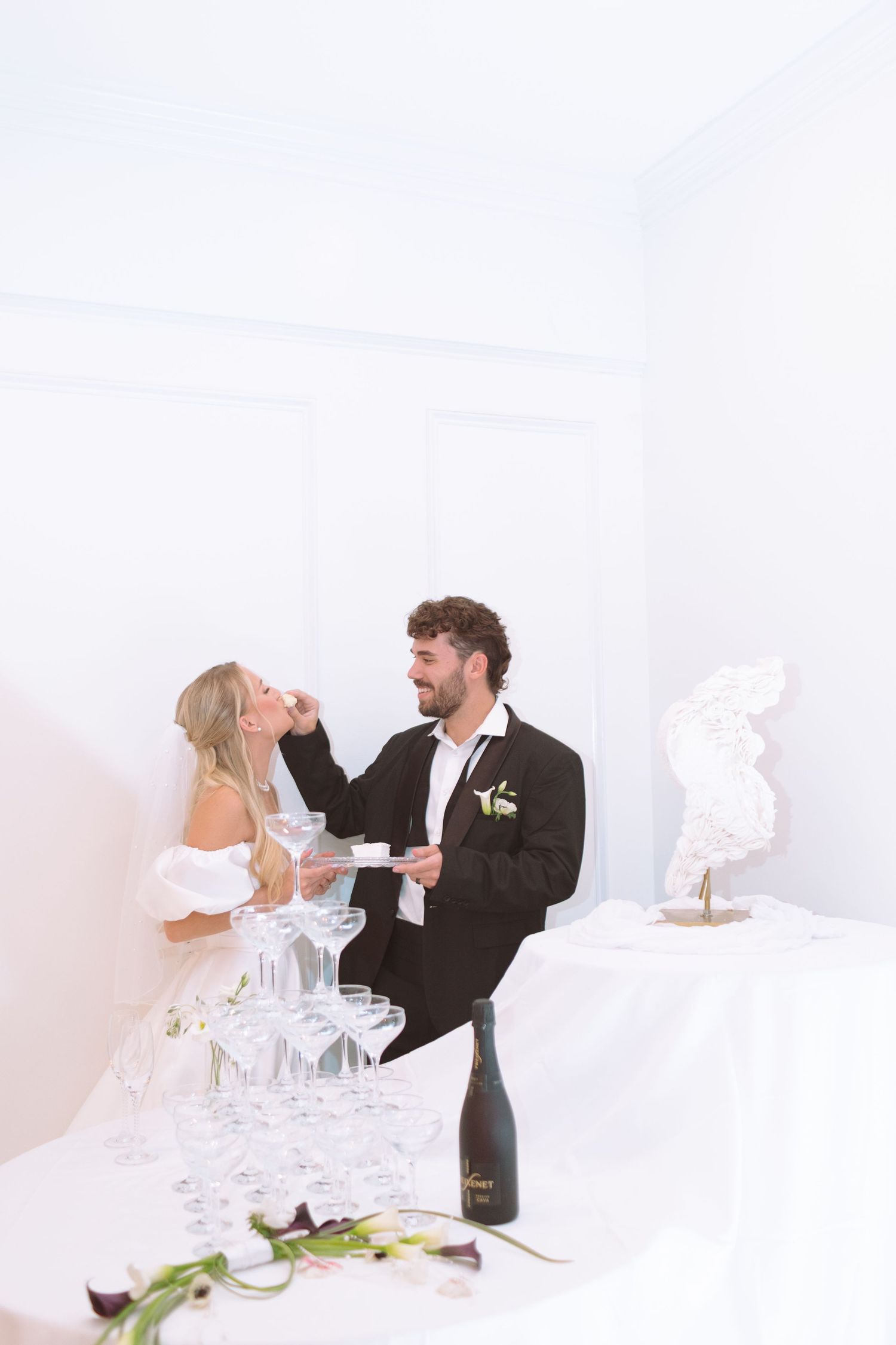 Groom feeding bride a piece of wedding cake near a champagne tower at their elegant white reception.
