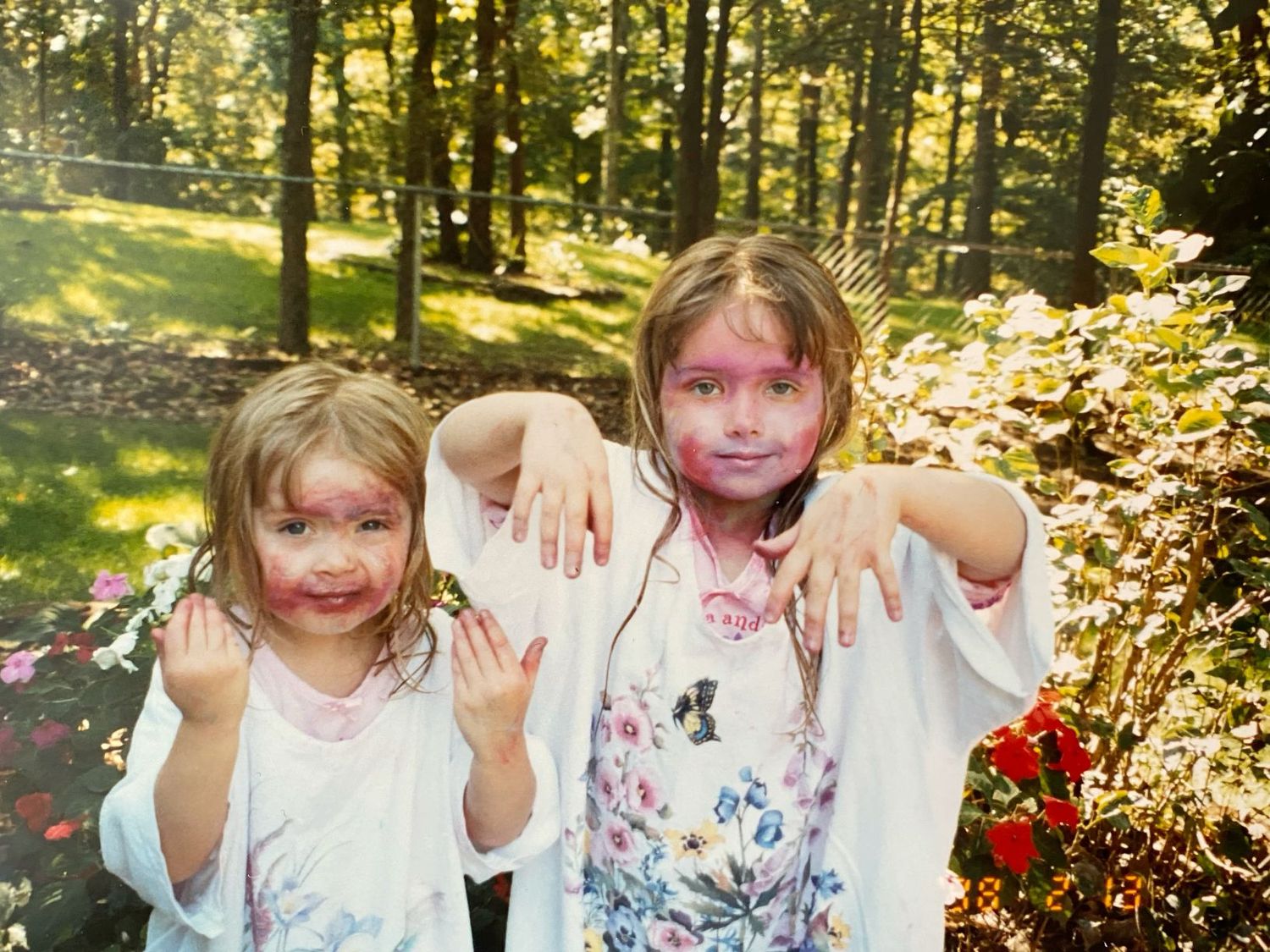 Two young girls covered in paint show their messy hands while playing outdoors.