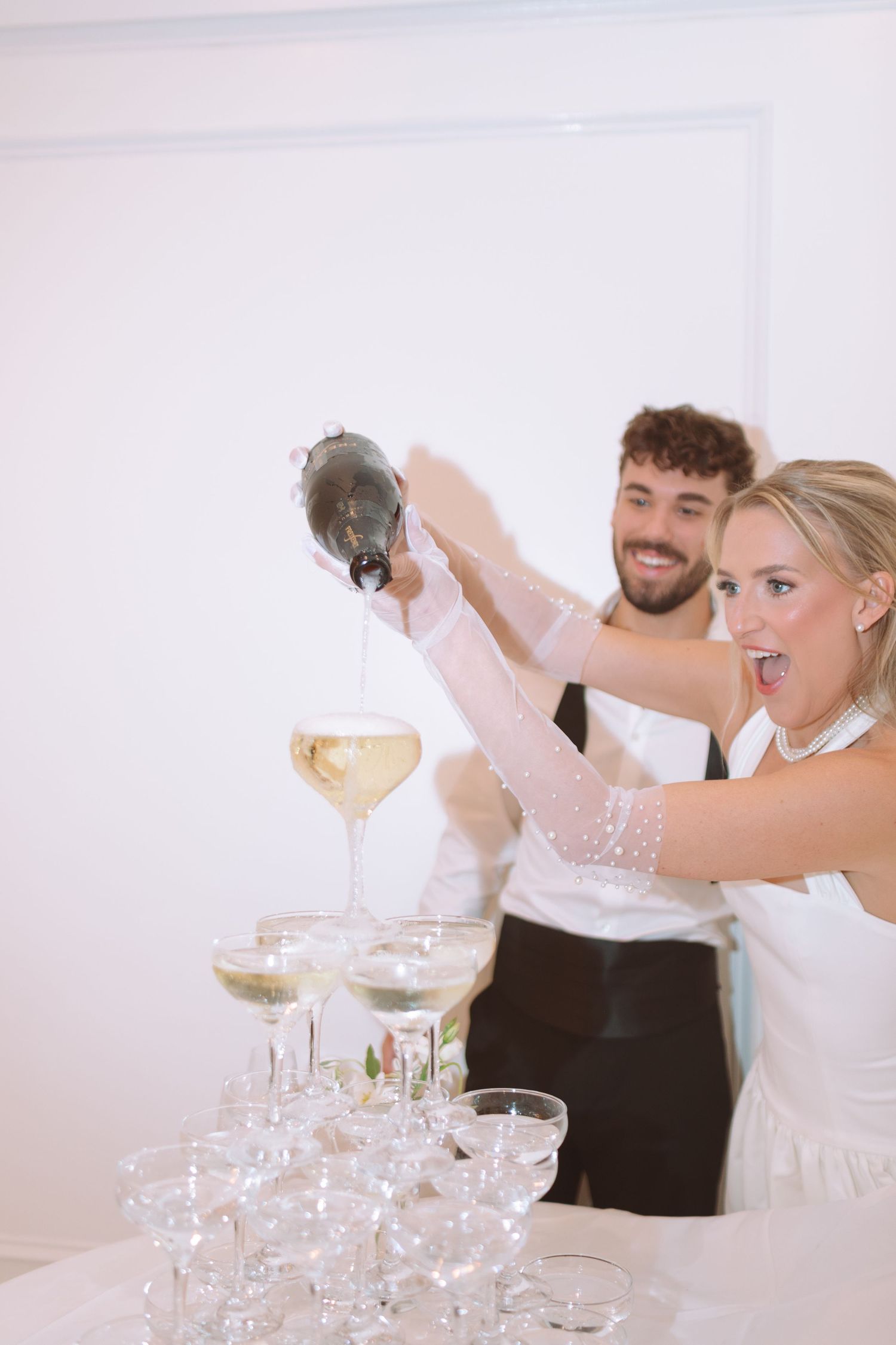 Excited bride and groom pouring champagne into a glass tower at their wedding celebration.