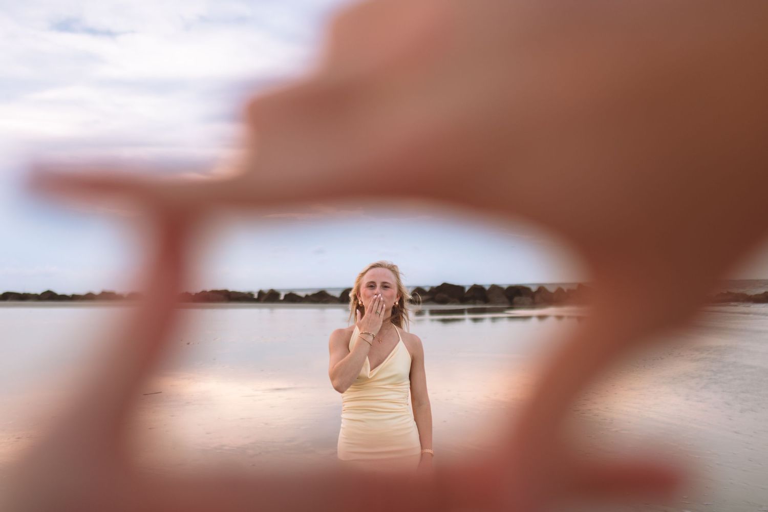 Blonde woman in white dress blowing a kiss framed by hands near a serene waterfront at sunset.