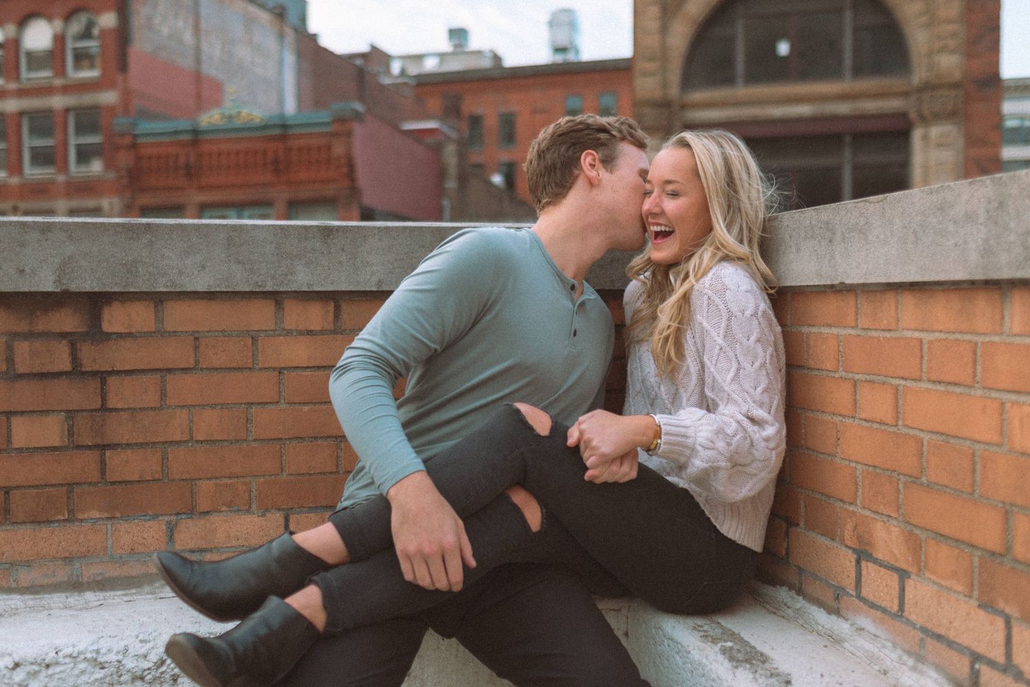 Laughing couple sitting on rooftop brick ledge in urban setting sharing a joyful moment.