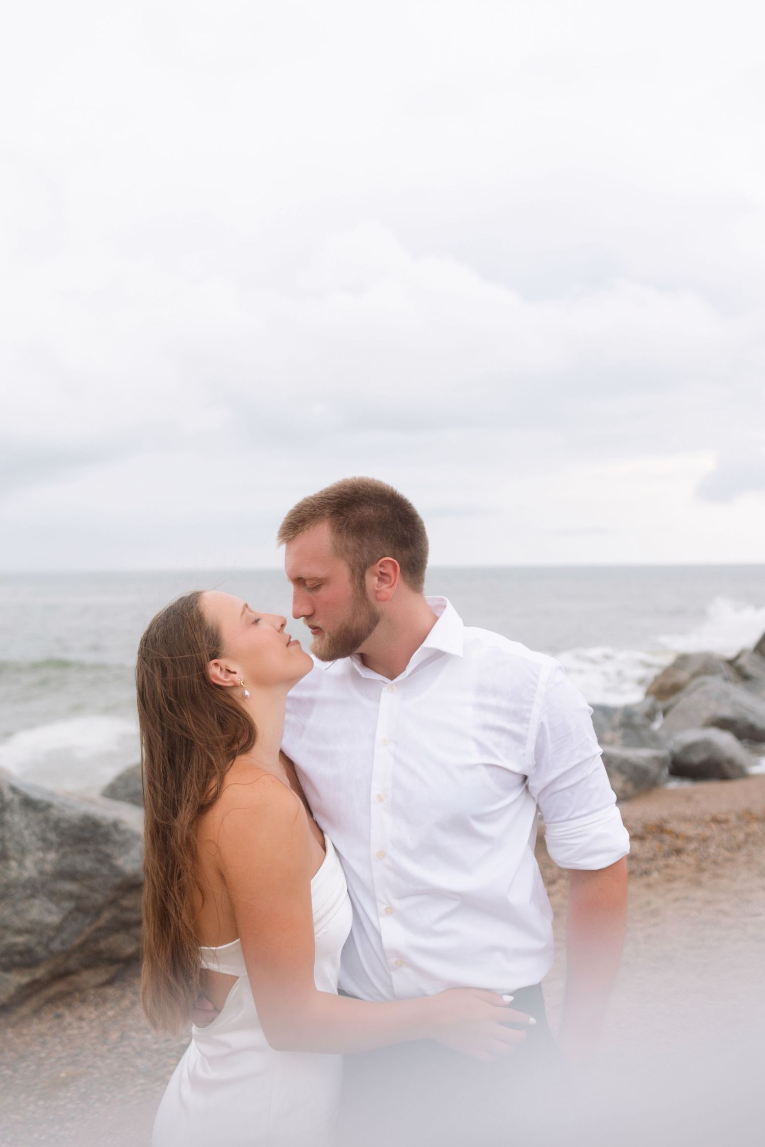 Romantic couple sharing a kiss on rocky beach shoreline, woman in white dress and man in white shirt.