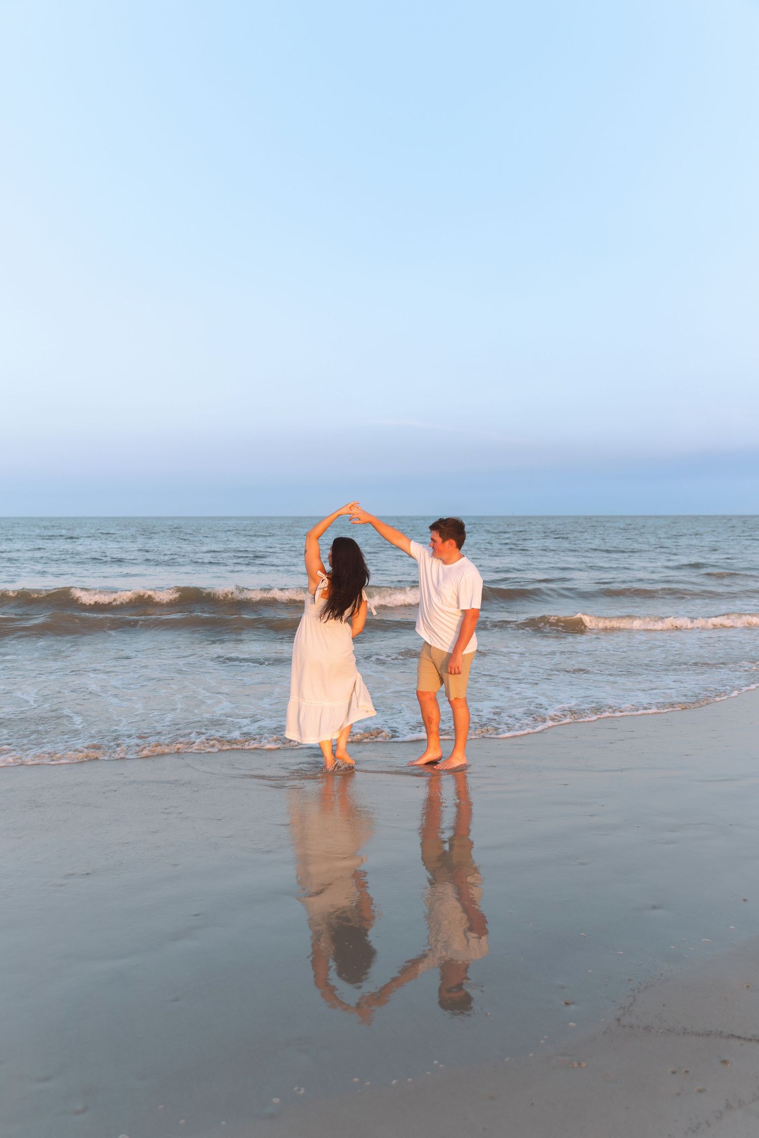 Couple dancing on the beach at sunset, reflecting on wet sand with ocean waves behind them.