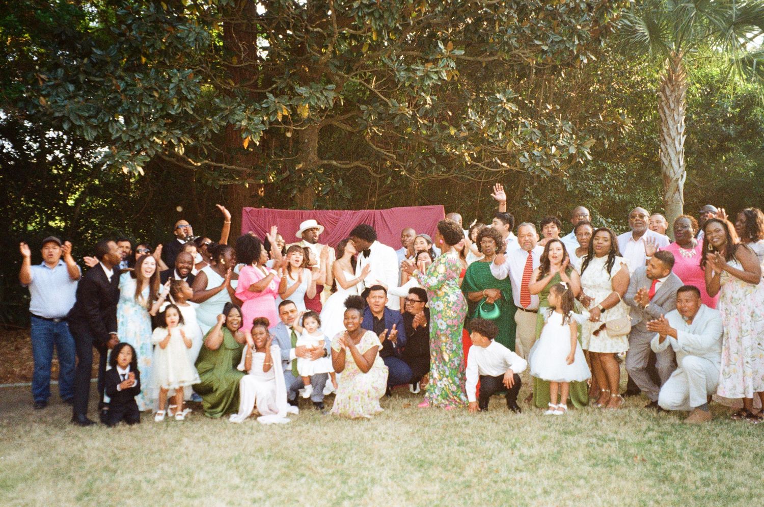 Large joyful wedding group photo with diverse guests celebrating outdoors under lush green trees in natural light.