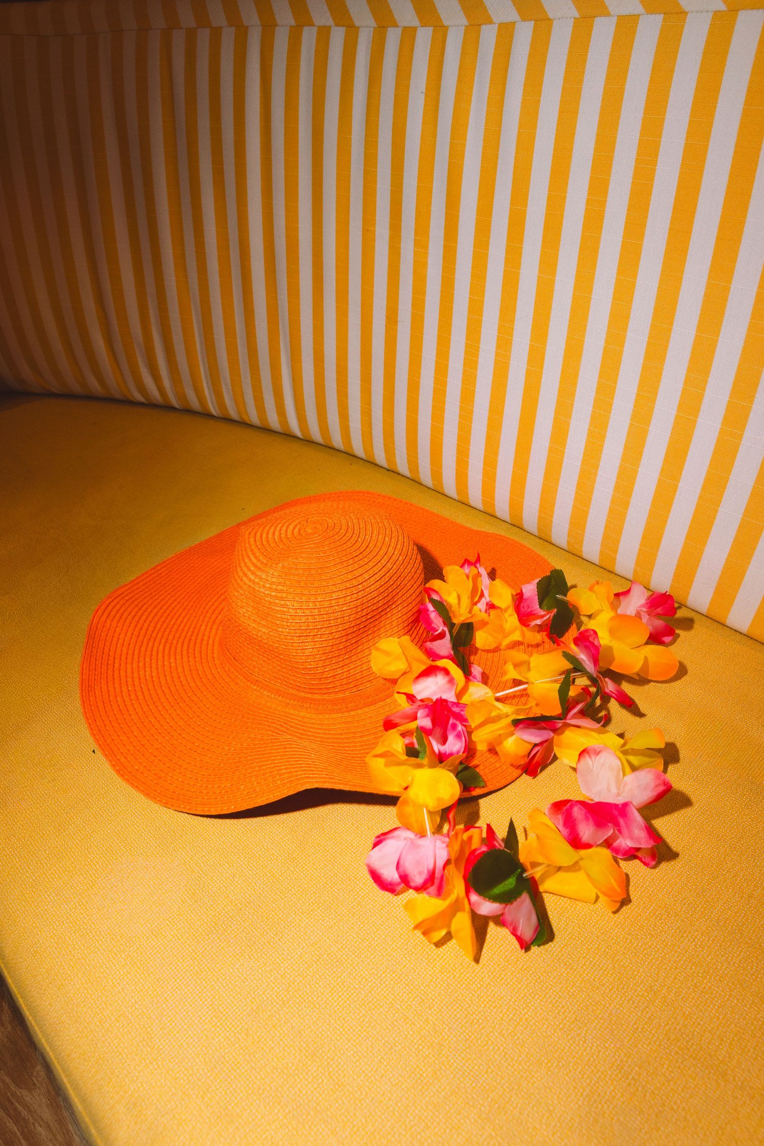 Orange wide-brim straw sun hat with colorful tropical flower lei on yellow surface with striped background.