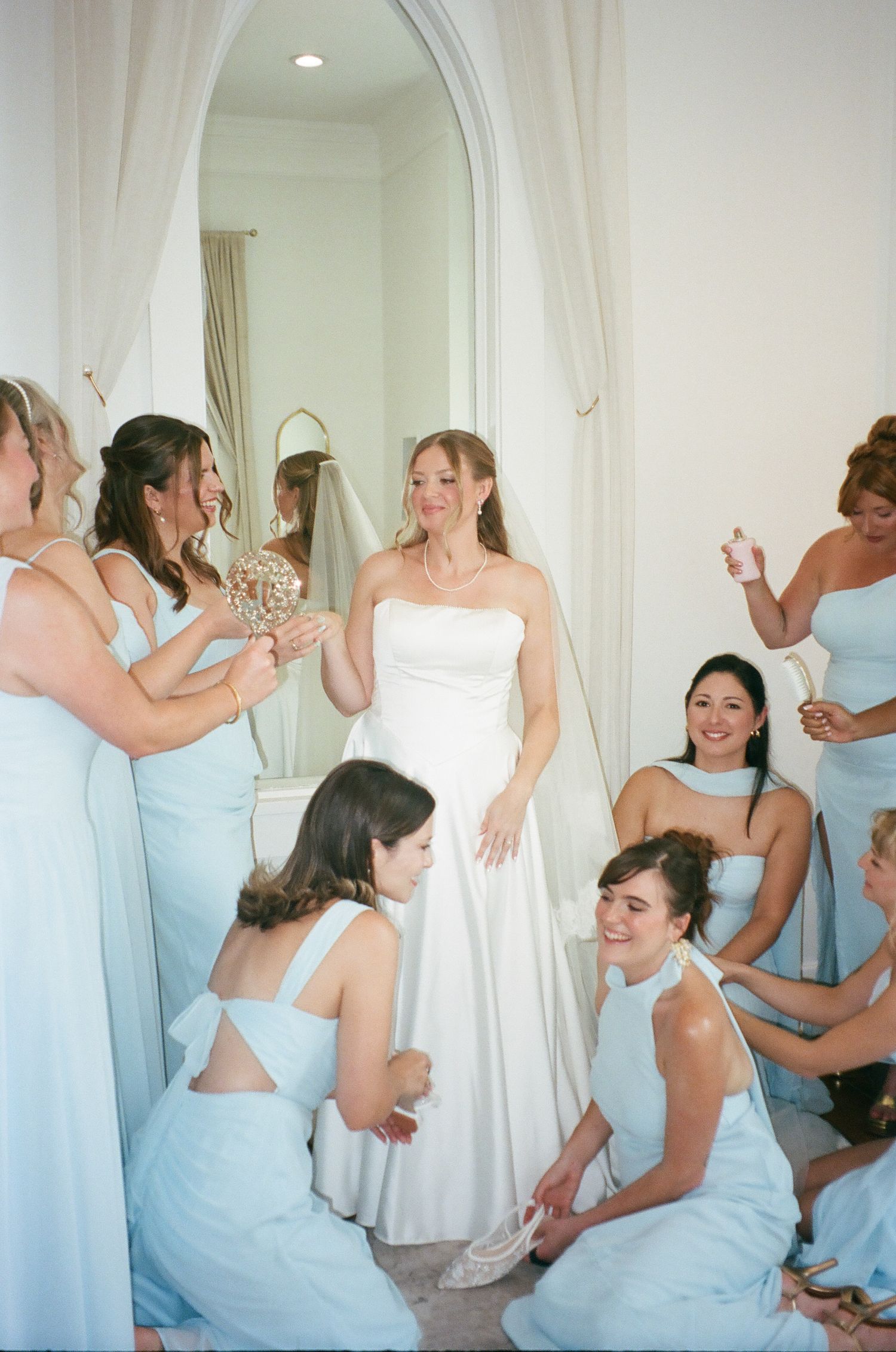 Bride in white gown surrounded by bridesmaids in light blue dresses helping her prepare in elegant room.