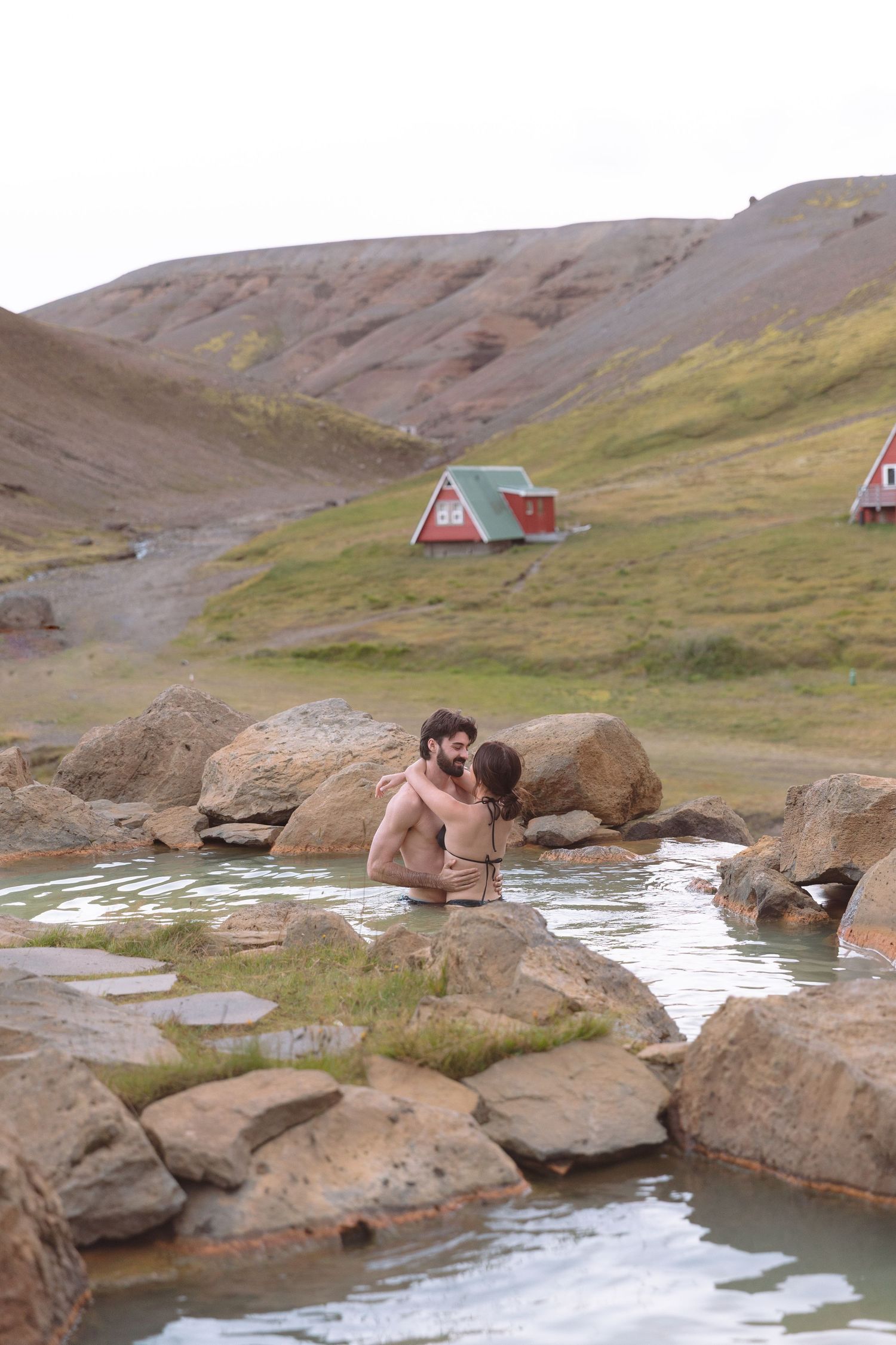 Romantic couple kissing in an Icelandic geothermal hot spring with red mountain huts in background.