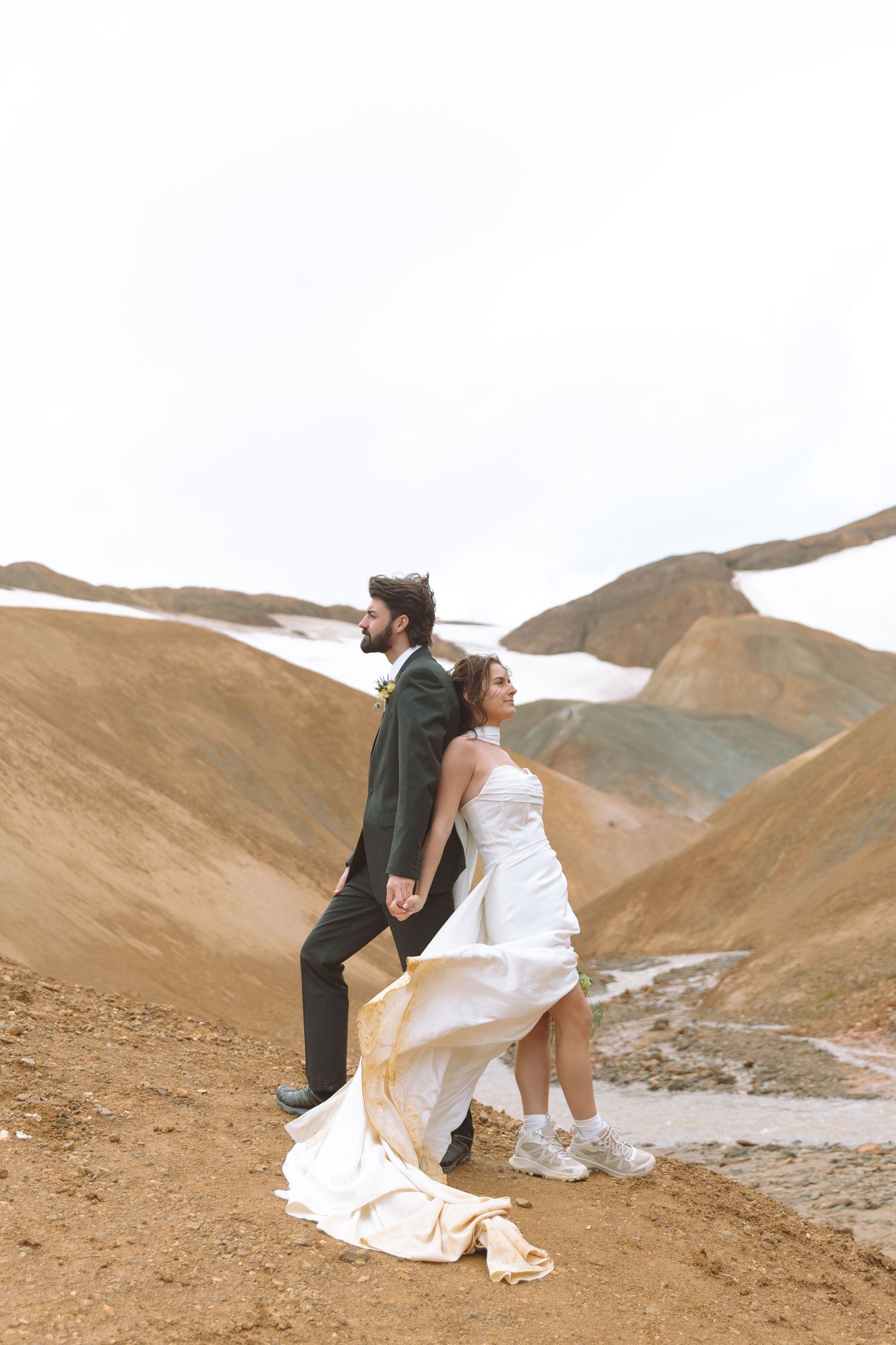 Bride and groom stand back to back on Iceland's golden volcanic hillside with snowy peaks in the distance.