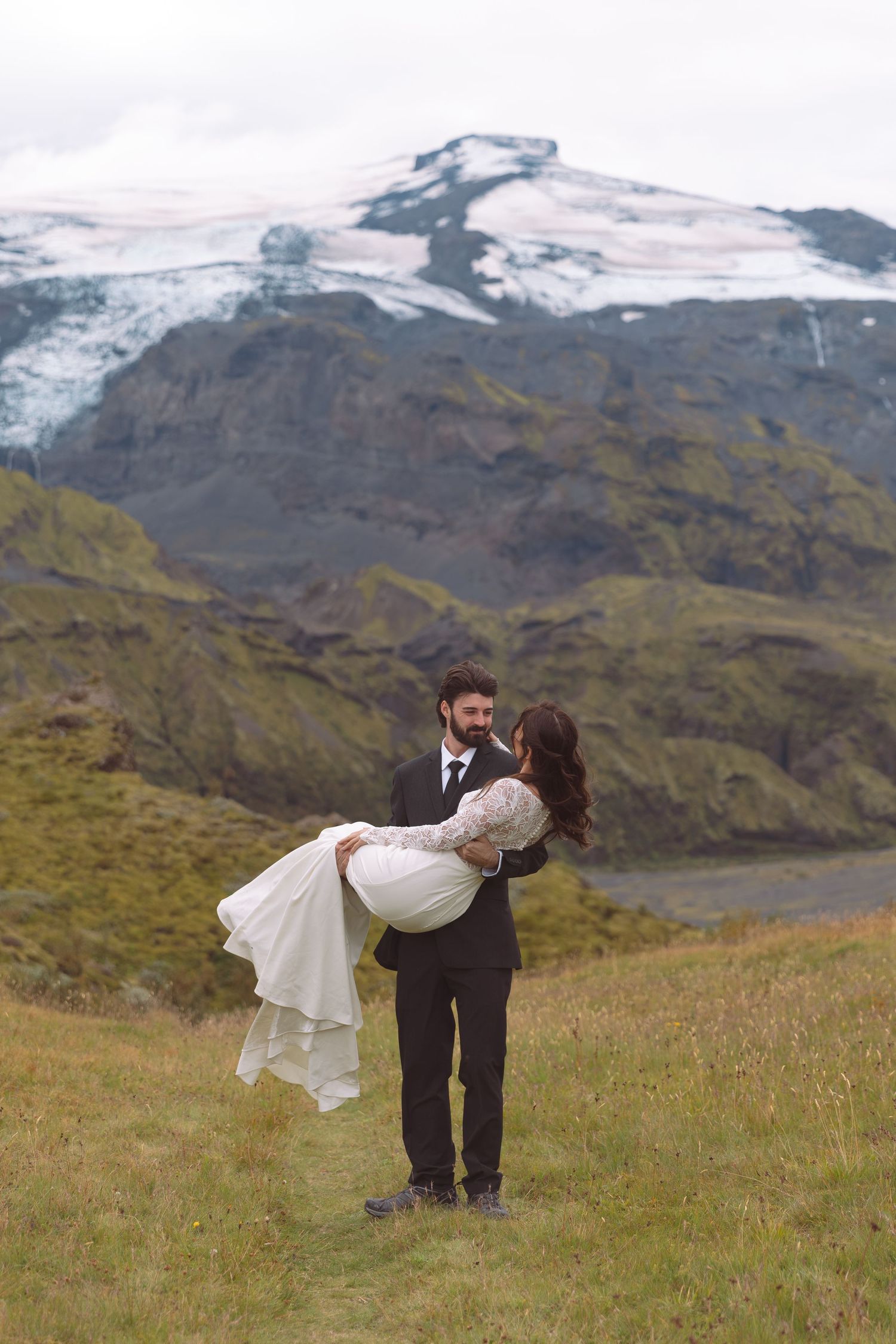 Groom carries smiling bride in white lace gown through a grassy meadow with snow-capped mountains behind.
