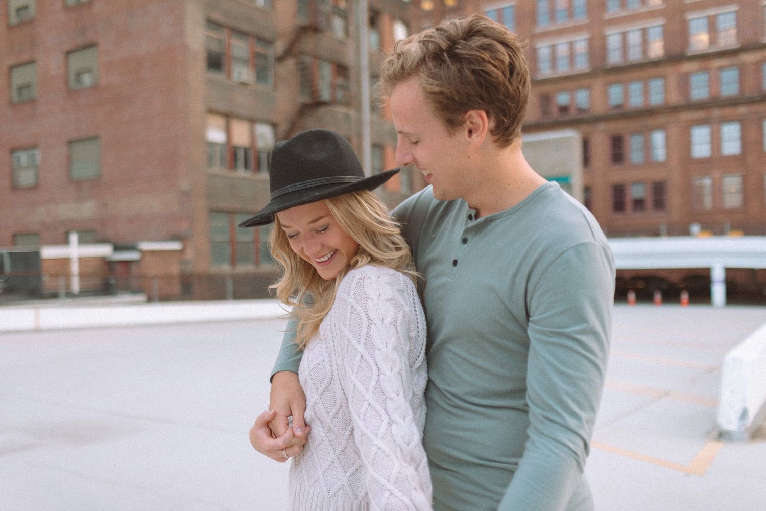 Smiling couple in casual attire embracing on a rooftop with brick buildings in background.