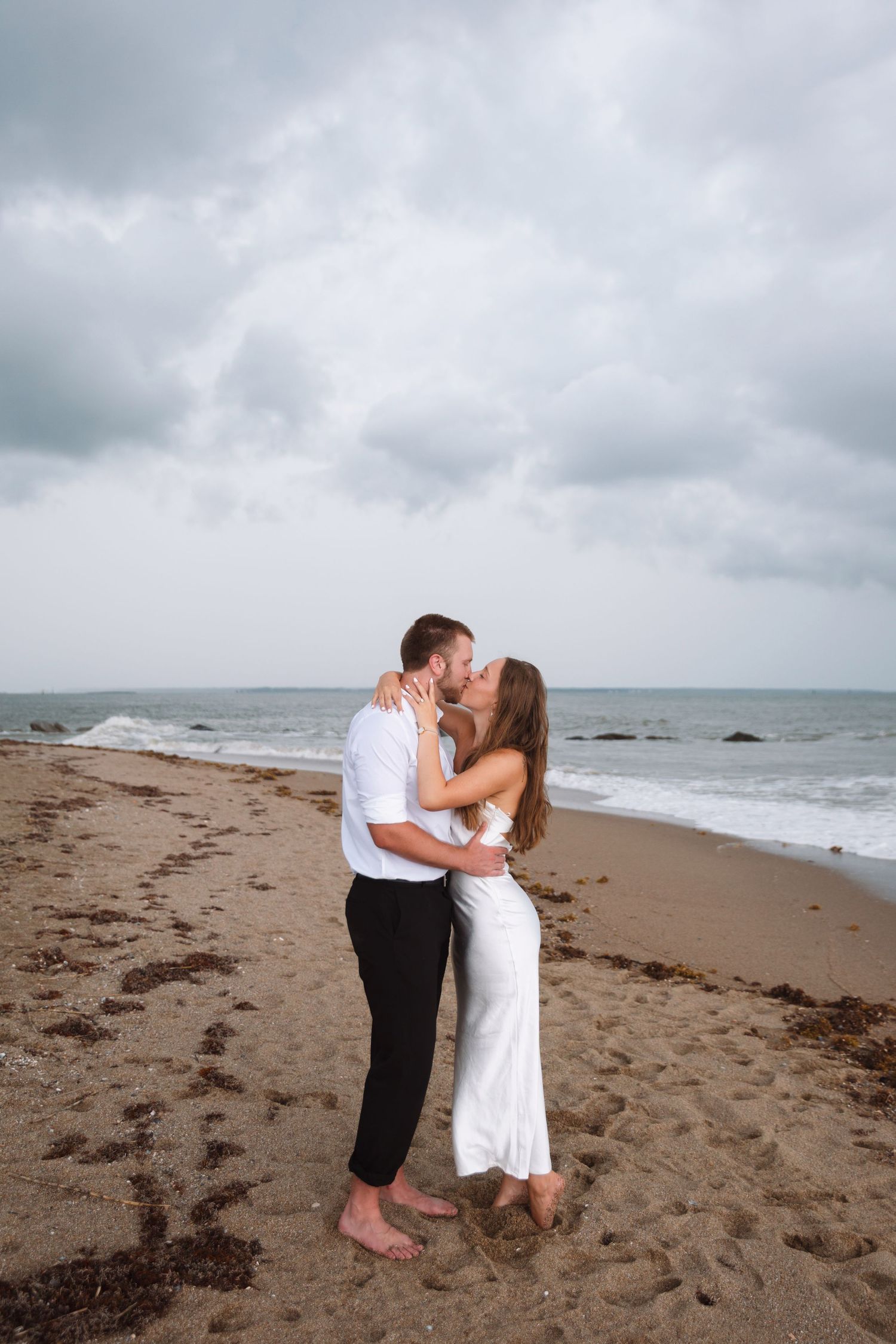 Couple sharing a kiss on a sandy beach in white outfits with dramatic cloudy skies and ocean waves behind them.