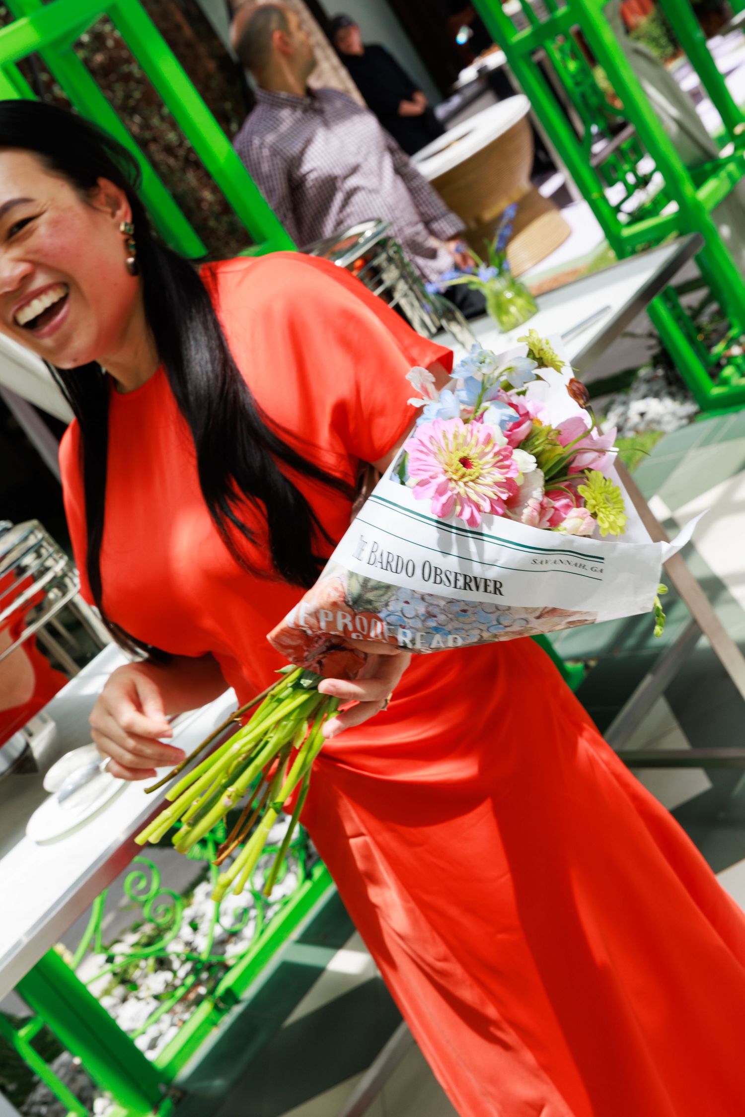 Laughing woman in red dress holding colorful flower bouquet wrapped in newspaper at an outdoor event.